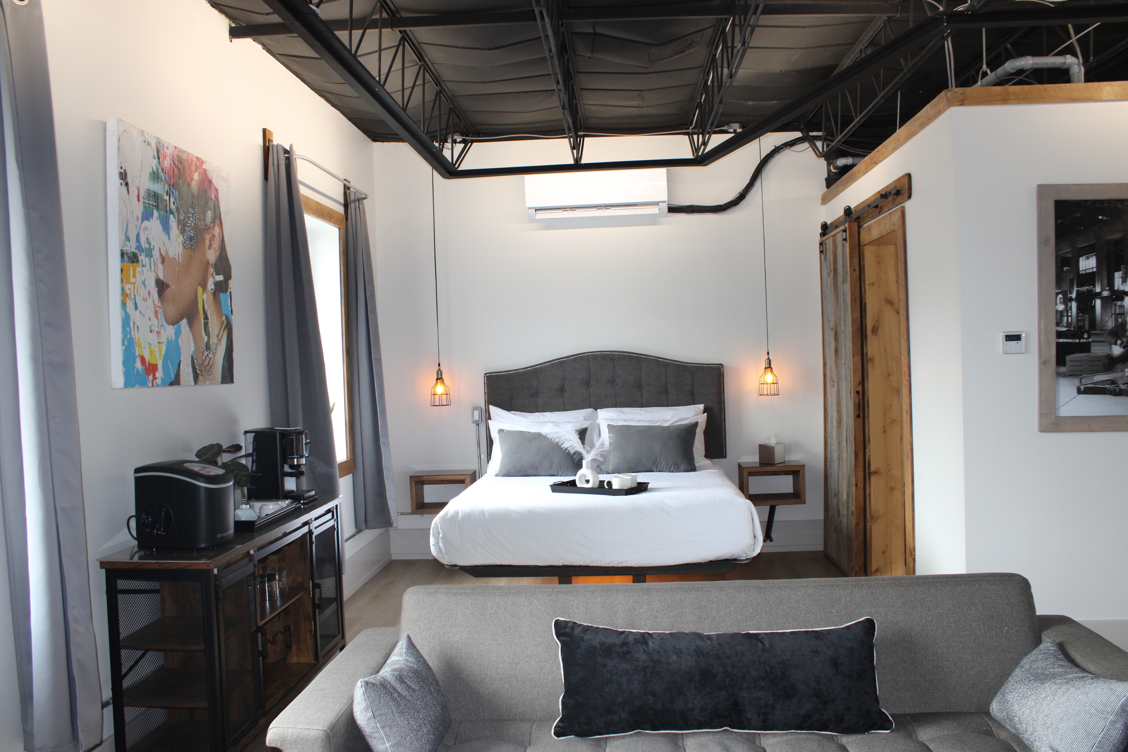 A trendy industrial loft bedroom with a dark, exposed ceiling, a queen bed with a tufted headboard, and vibrant modern artwork.