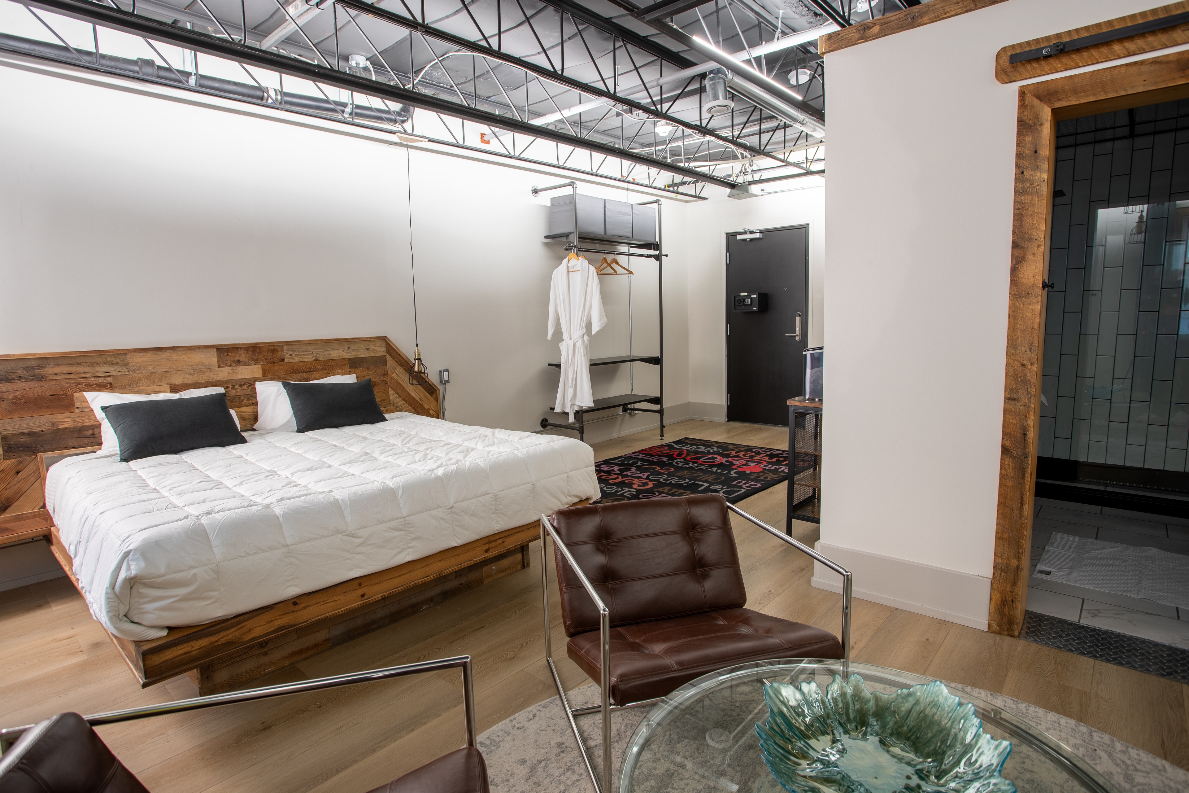 A bright, modern industrial loft bedroom with a reclaimed wood bed, leather chairs, open closet storage, and a view into the shower.