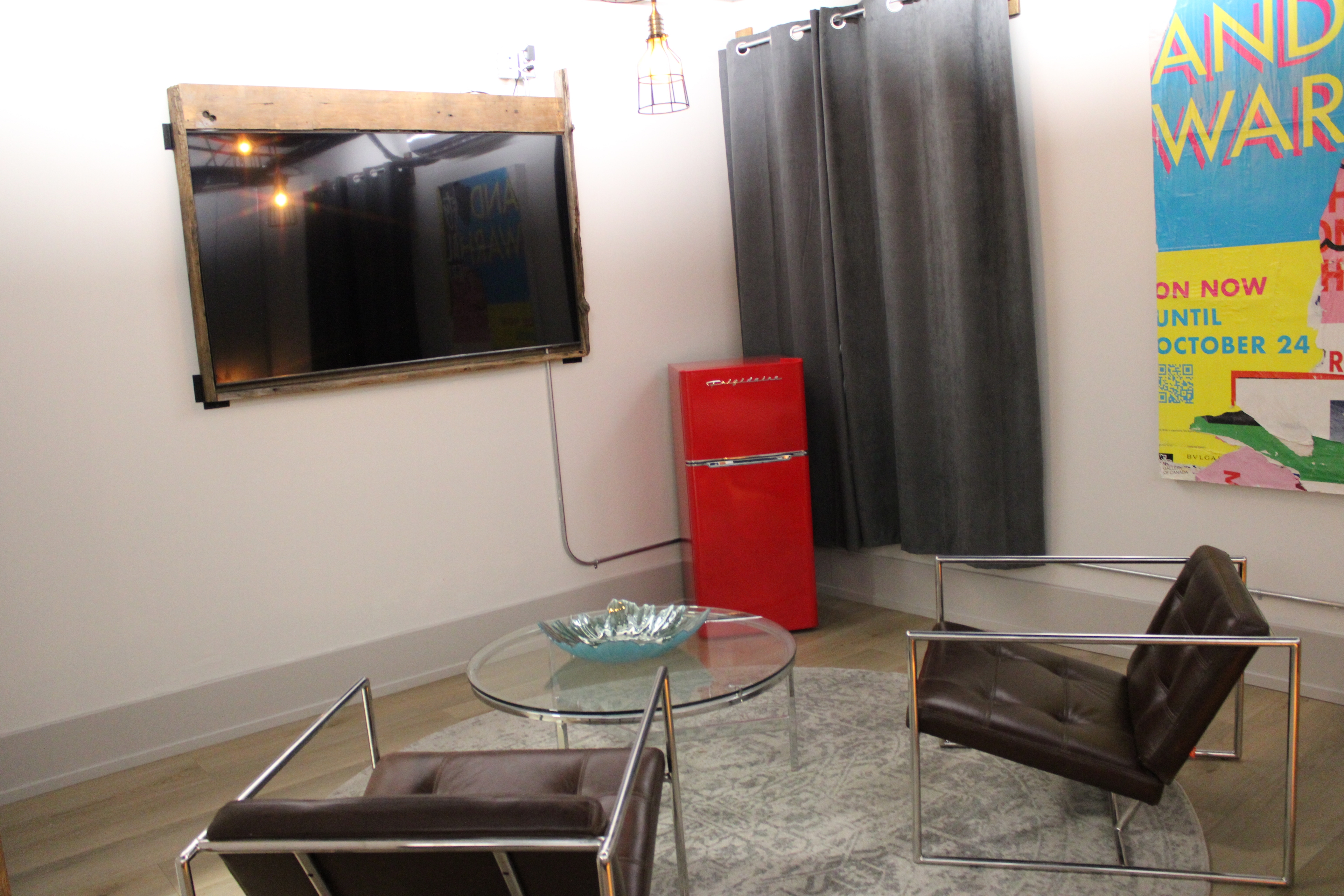 A stylish modern lounge area with dark leather and chrome chairs, a red retro mini-fridge, a wall-mounted TV, and pop art on the wall.