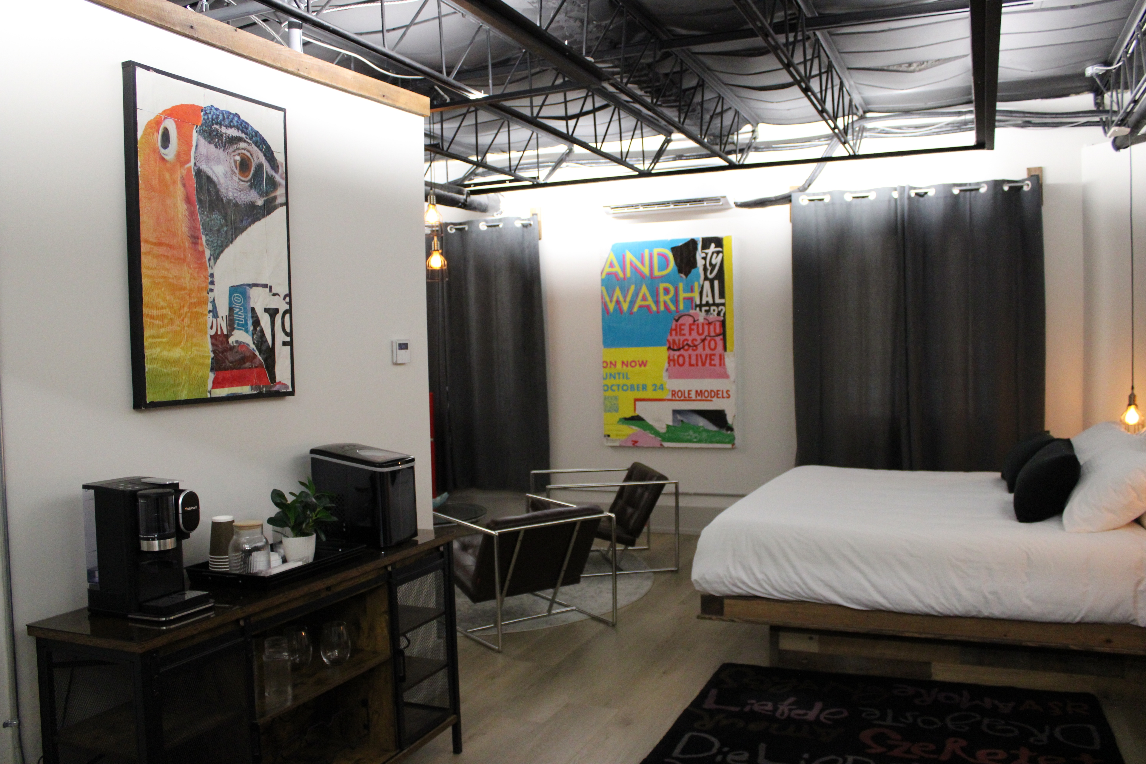 A stylish industrial-loft room with a black ceiling, modern wood bed, vibrant pop art, and a coffee station on a dark wood cabinet.