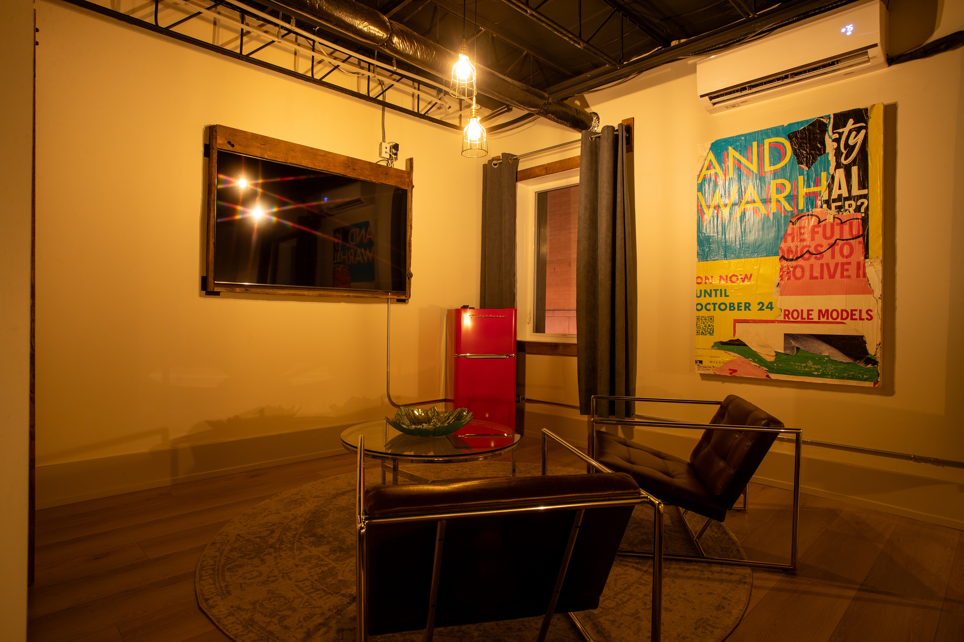 A cozy, moody industrial-style lounge with exposed ceiling, vintage leather chairs, a red mini-fridge, and vibrant pop art on the wall.