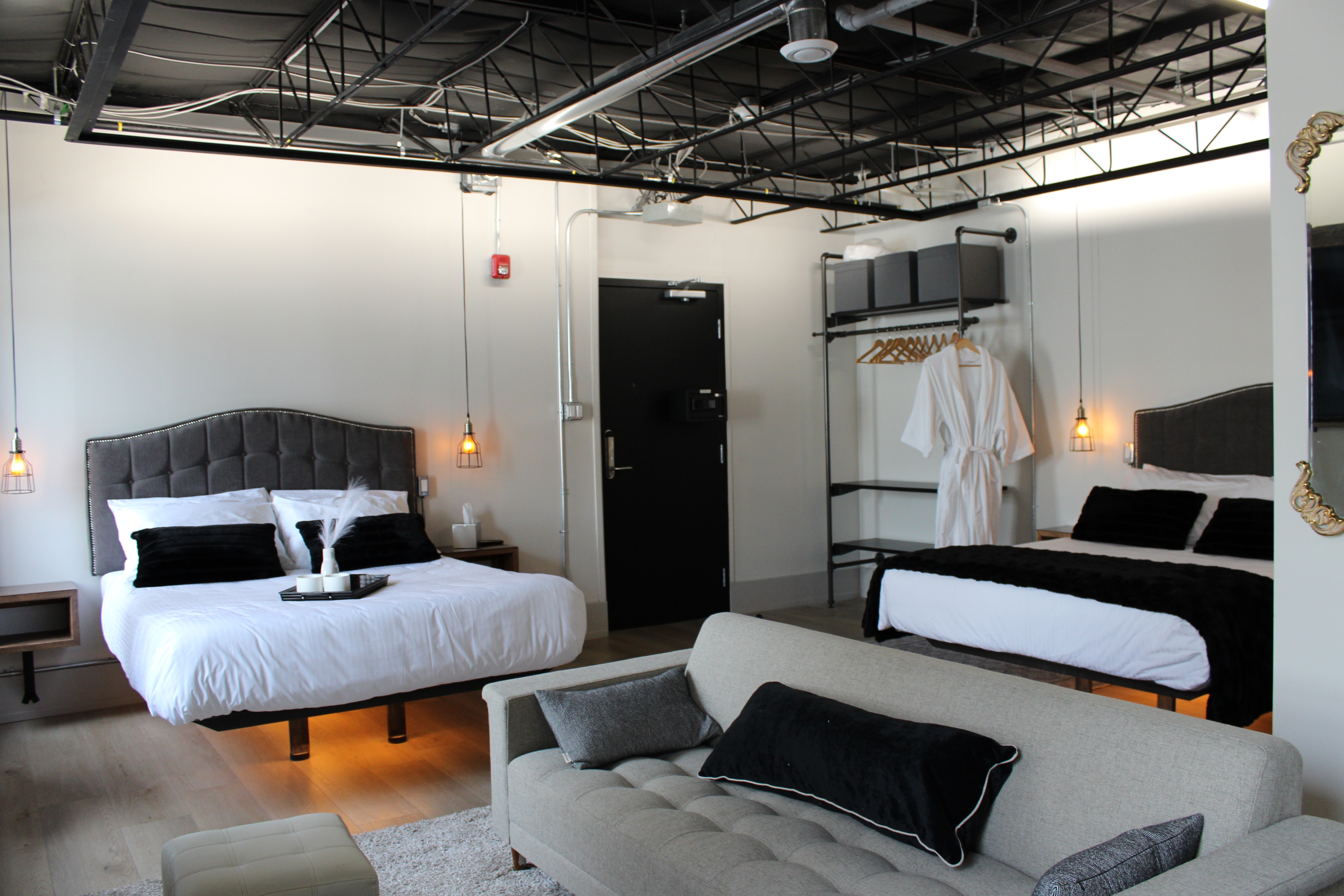 A modern industrial-style loft room featuring two queen beds with tufted headboards, a light gray sofa, and a black open ceiling.