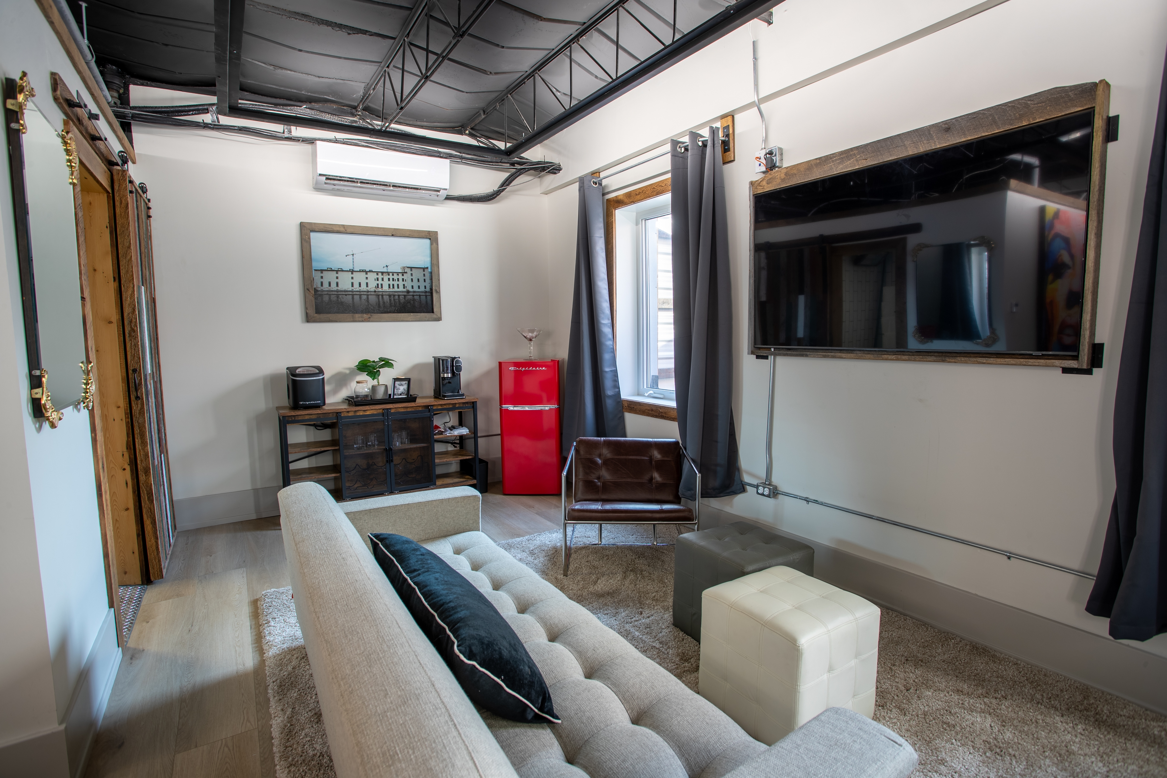 A trendy industrial loft living area with a light gray sofa, a large mounted TV, and a red retro mini-fridge next to a console table.