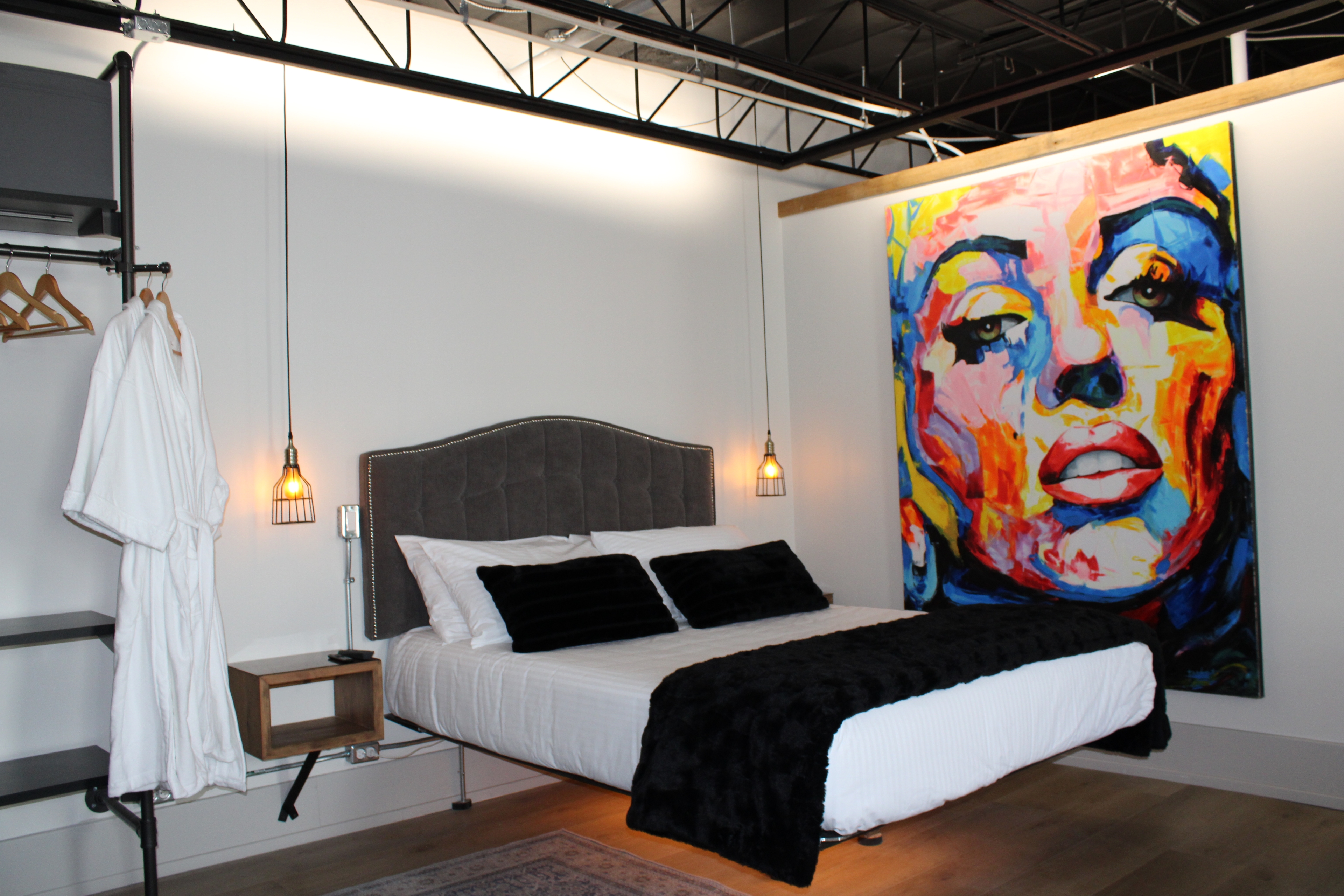 A stylish industrial-loft bedroom with a queen bed, a large, vibrant Marilyn Monroe pop art piece, and a robe hanging on an open closet.
