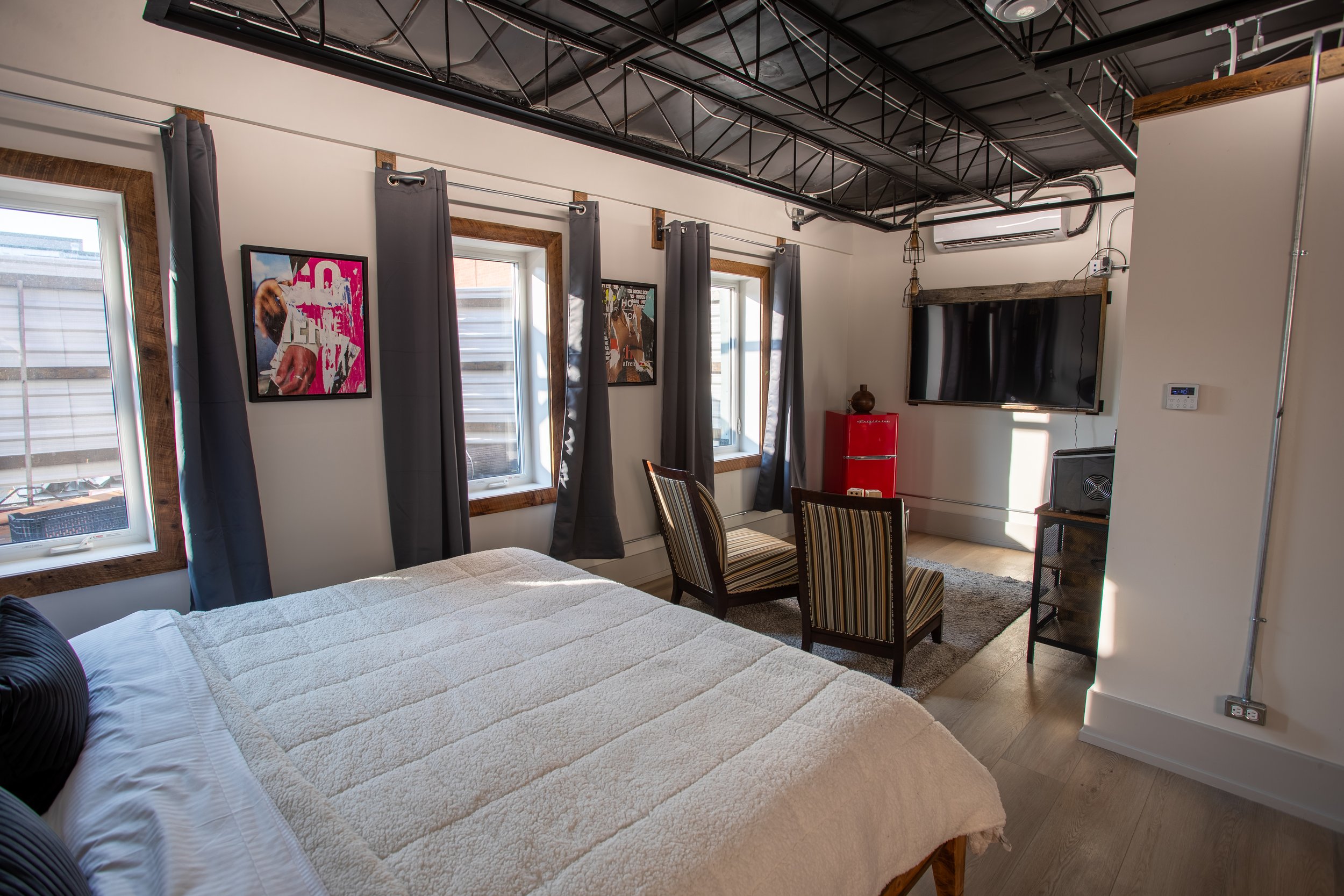 A bright, modern hotel room with a king bed, exposed black ceiling, multiple windows, a seating area, and pop art on the walls.