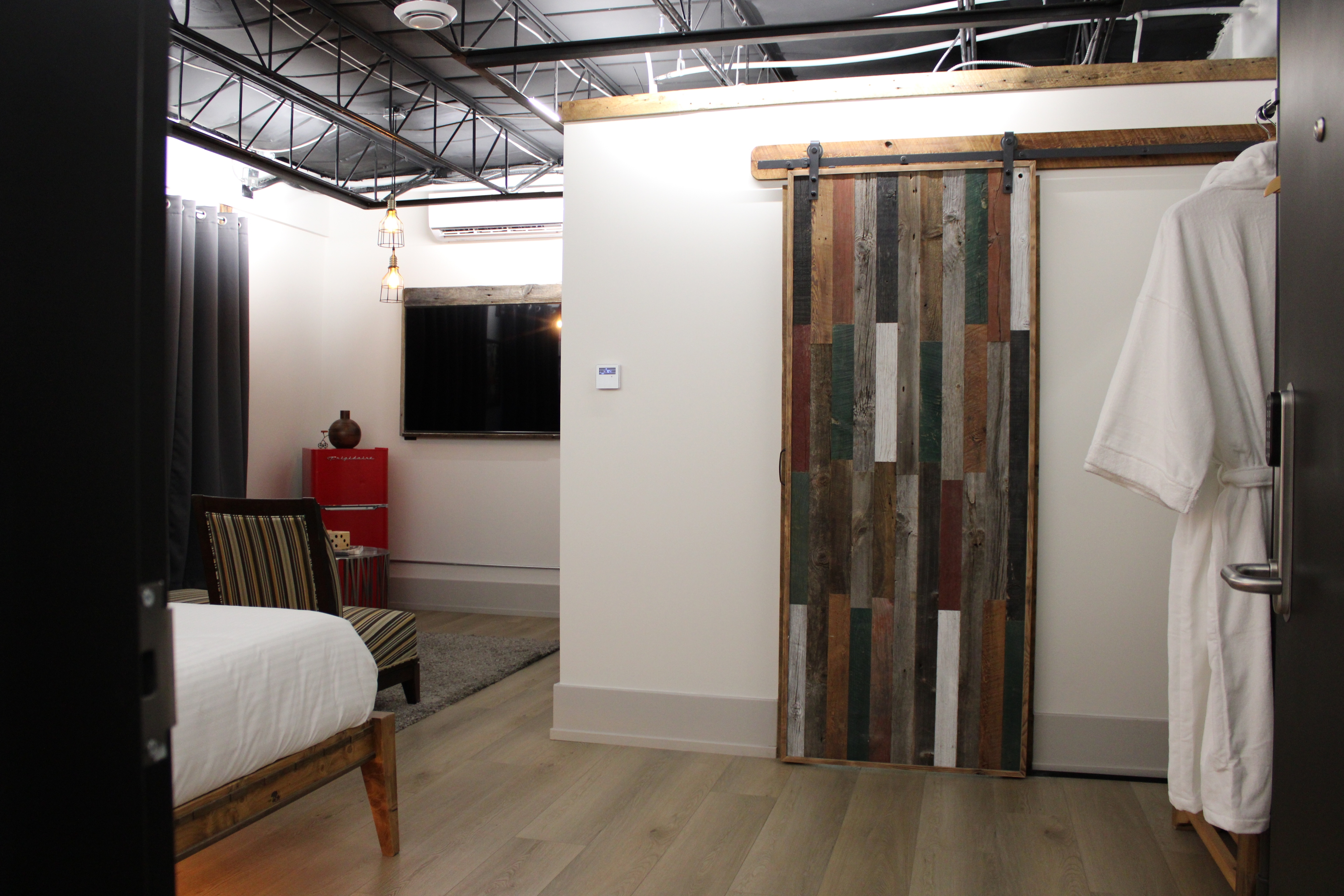 A stylish industrial-loft room with a rustic reclaimed wood sliding barn door, a plush white robe hanging up, and a view into the lounge area.