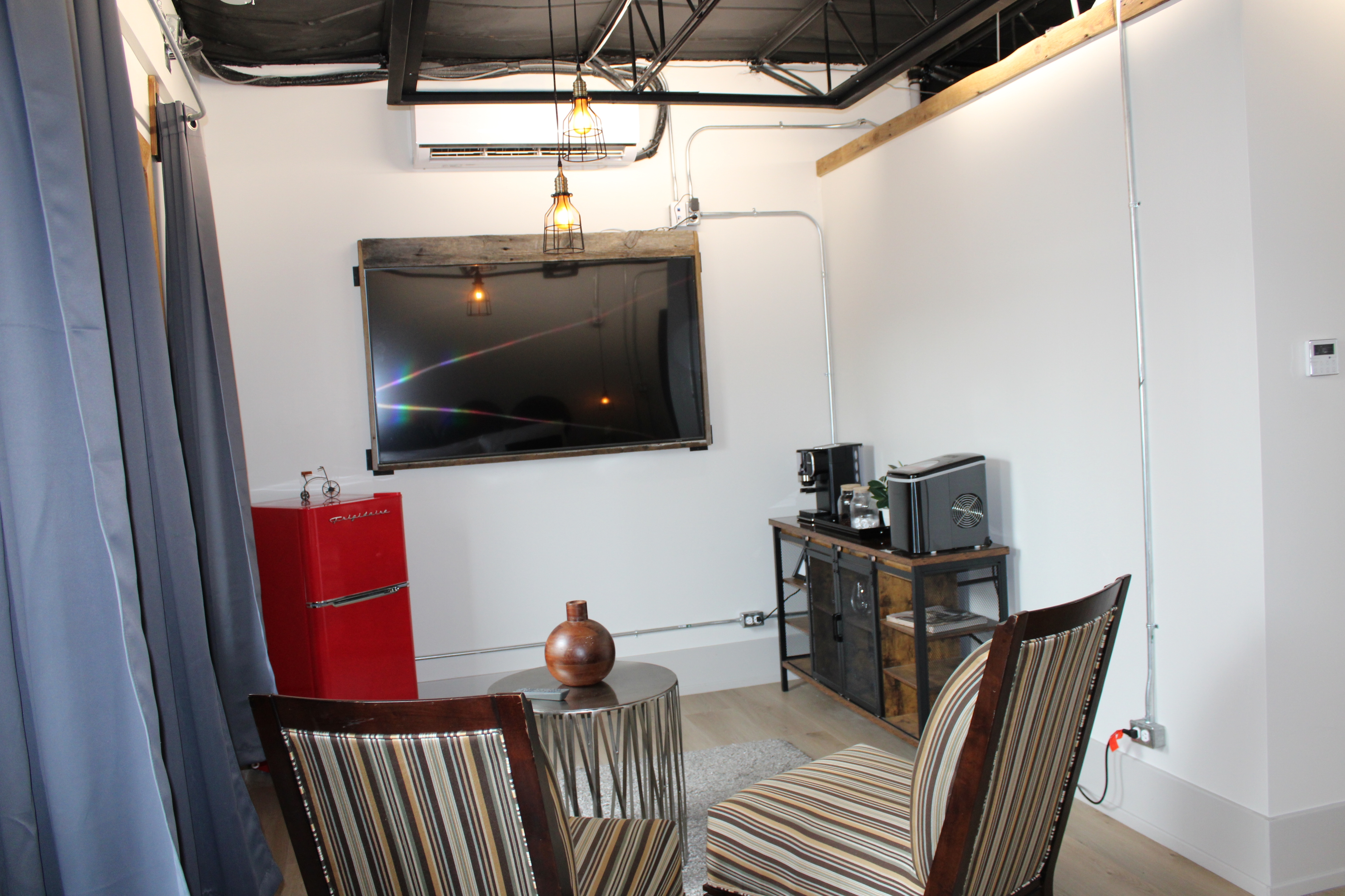 A small, trendy industrial lounge area with a mounted TV, two striped chairs, a red retro mini-fridge, and a coffee station.