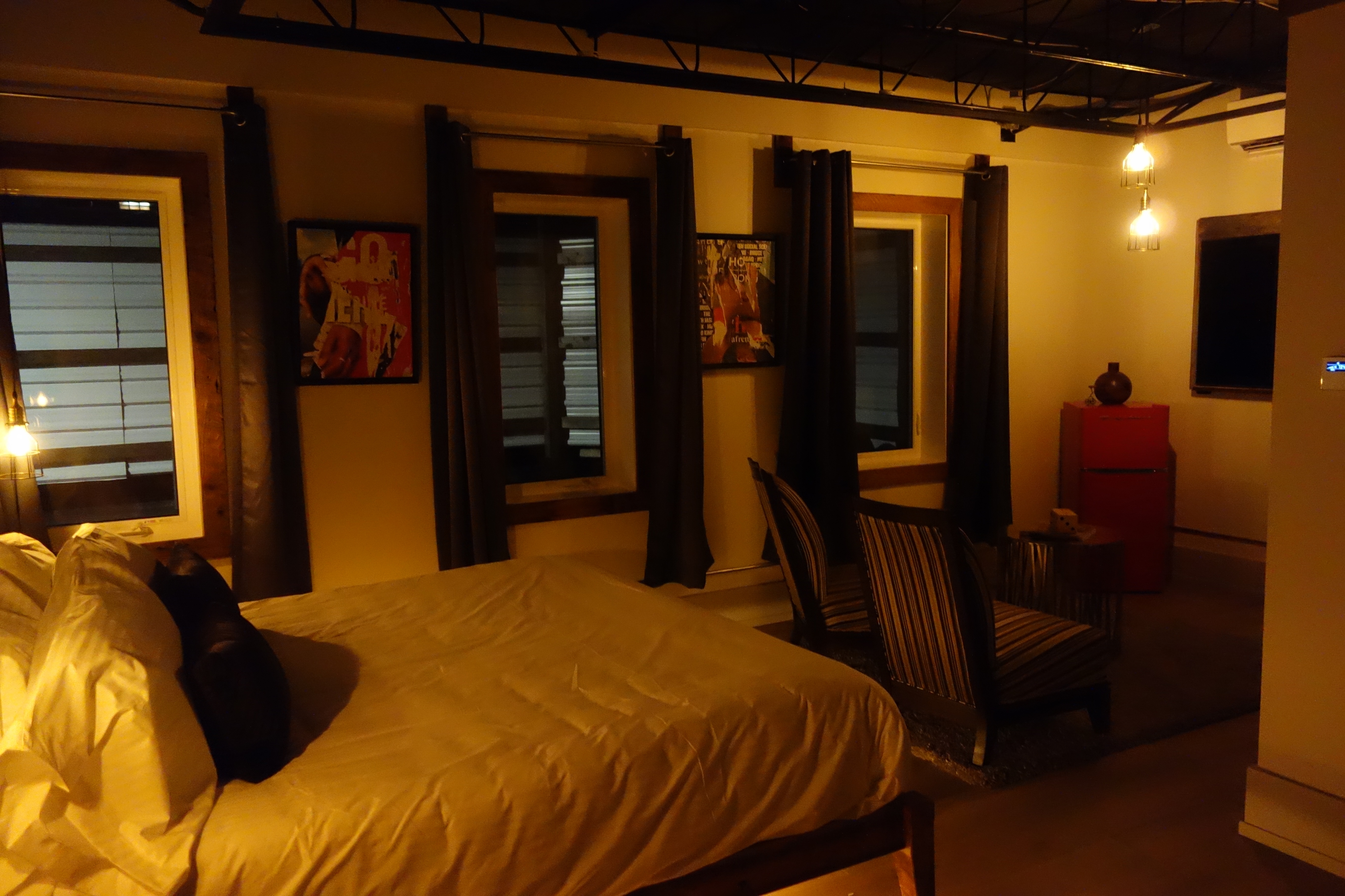 A dark, moody industrial loft bedroom with warm lighting, a king bed, black exposed ceiling, and window views into the city at night.