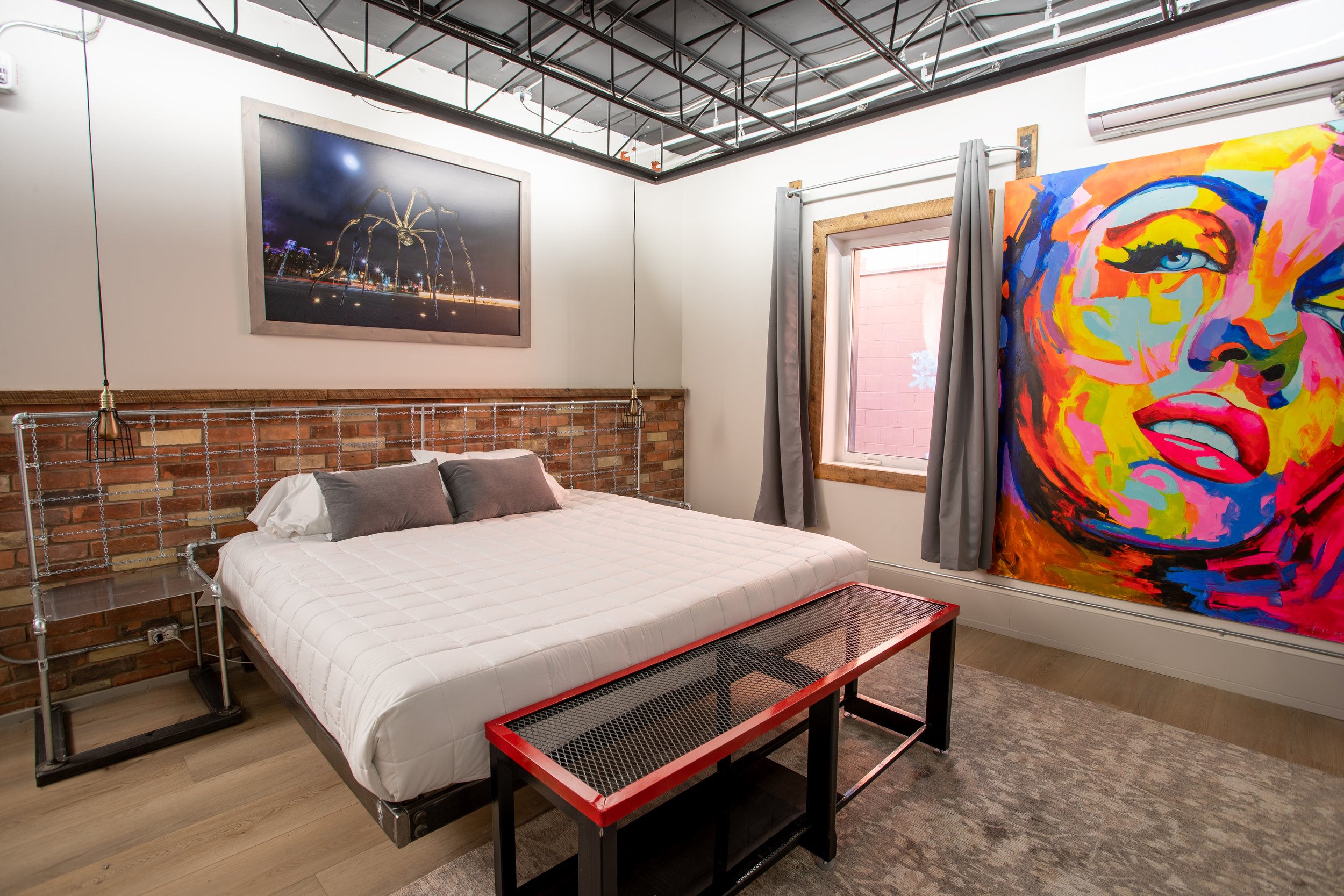 A bold, industrial loft bedroom featuring a king bed, brick wall detail, and a large, vibrant pop art canvas.