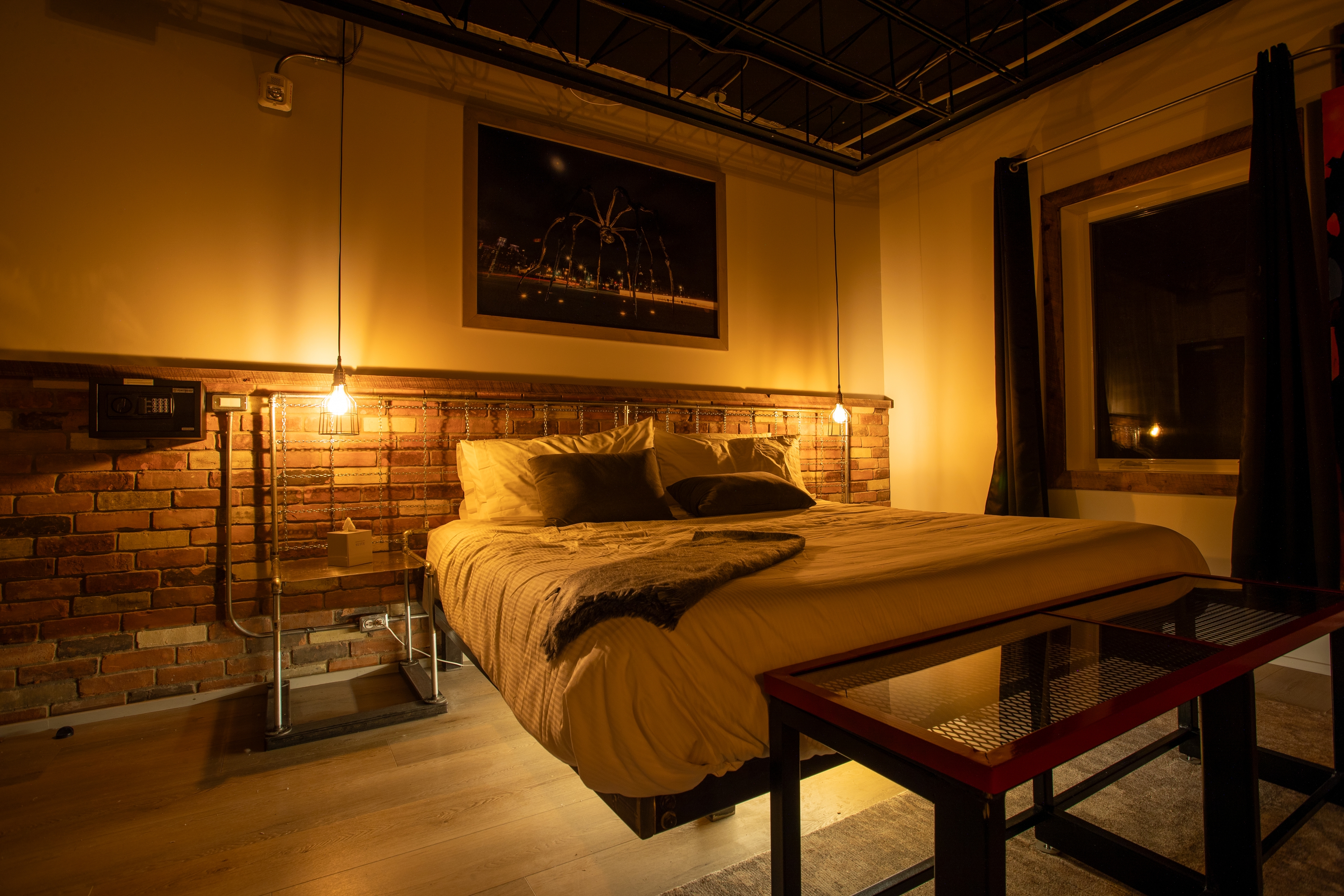 A moody, industrial loft bedroom featuring a brick accent wall, a low-profile bed with under-lighting, and warm hanging Edison bulb lights.
