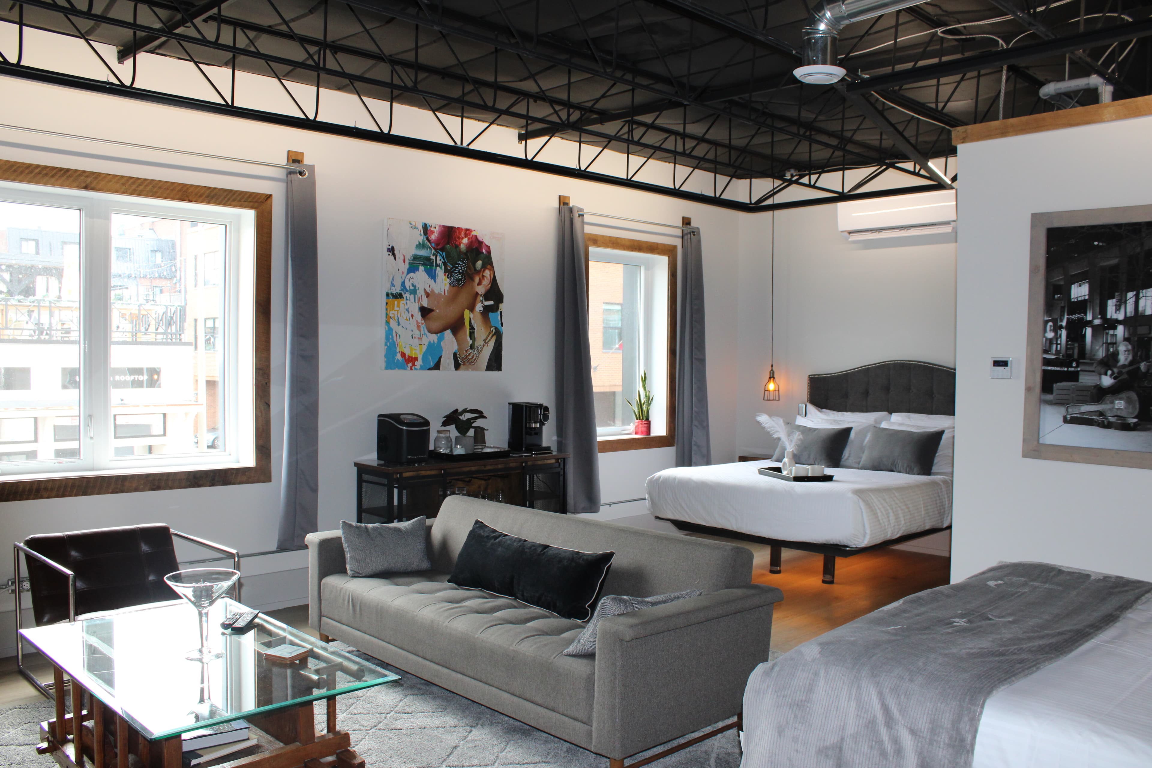 A stylish, open-concept loft space with a modern, industrial feel, featuring two beds, a gray sofa, high ceilings, and bright windows.