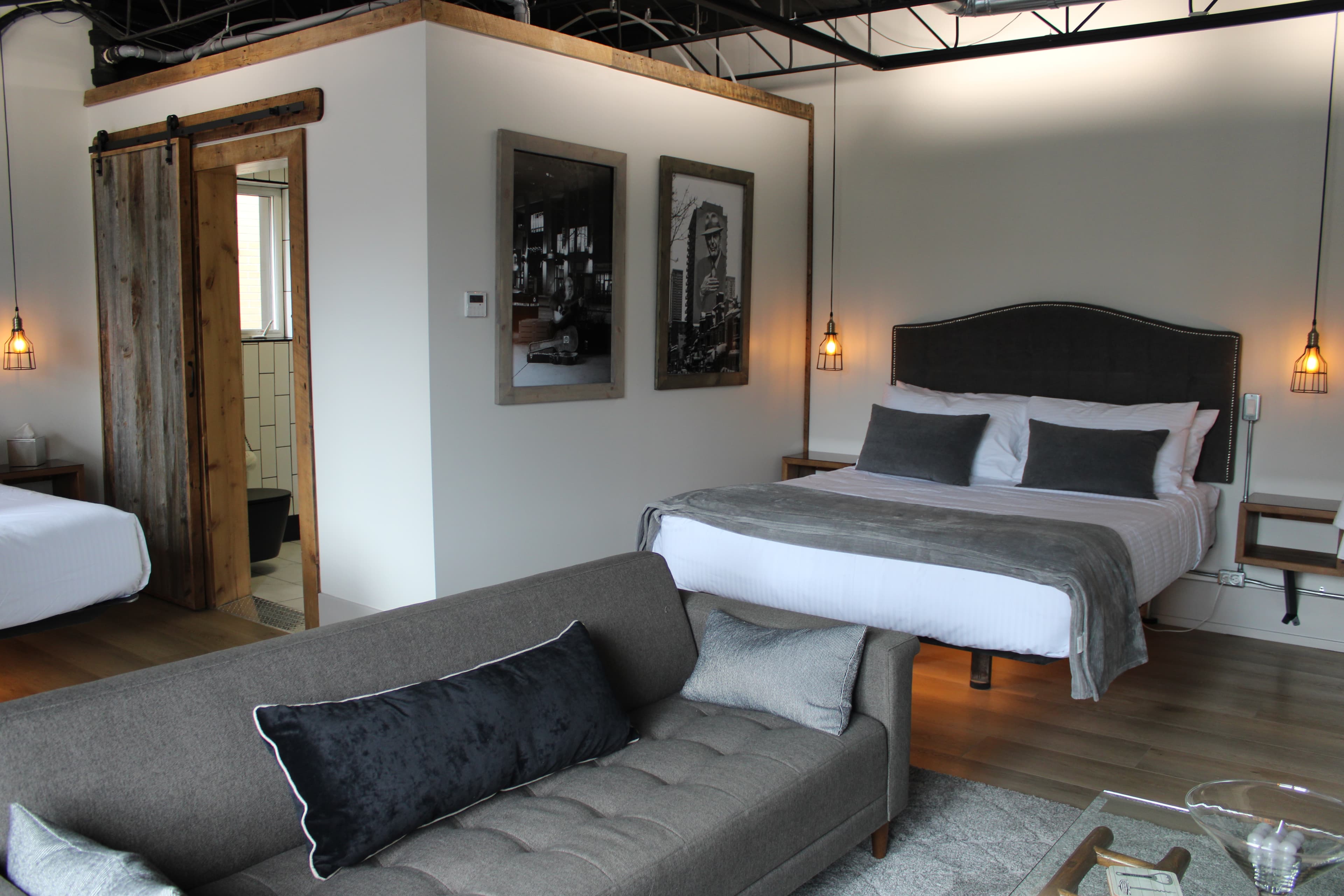 A spacious, modern industrial loft with two beds, a gray sofa, and a rustic sliding barn door leading to a bathroom.