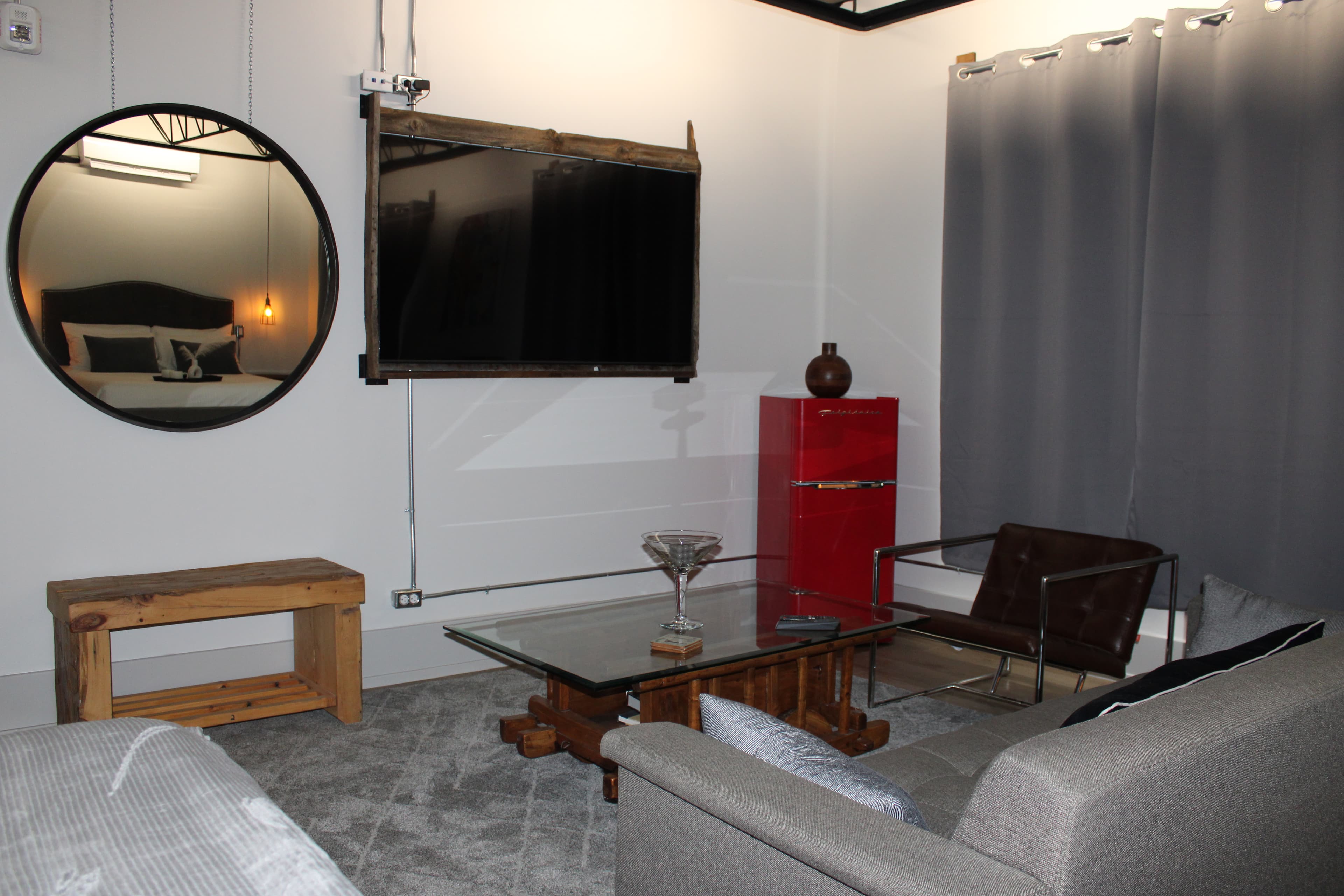 A stylish industrial living space with a gray sofa, glass coffee table, mounted TV, red retro mini-fridge, and a large round mirror.