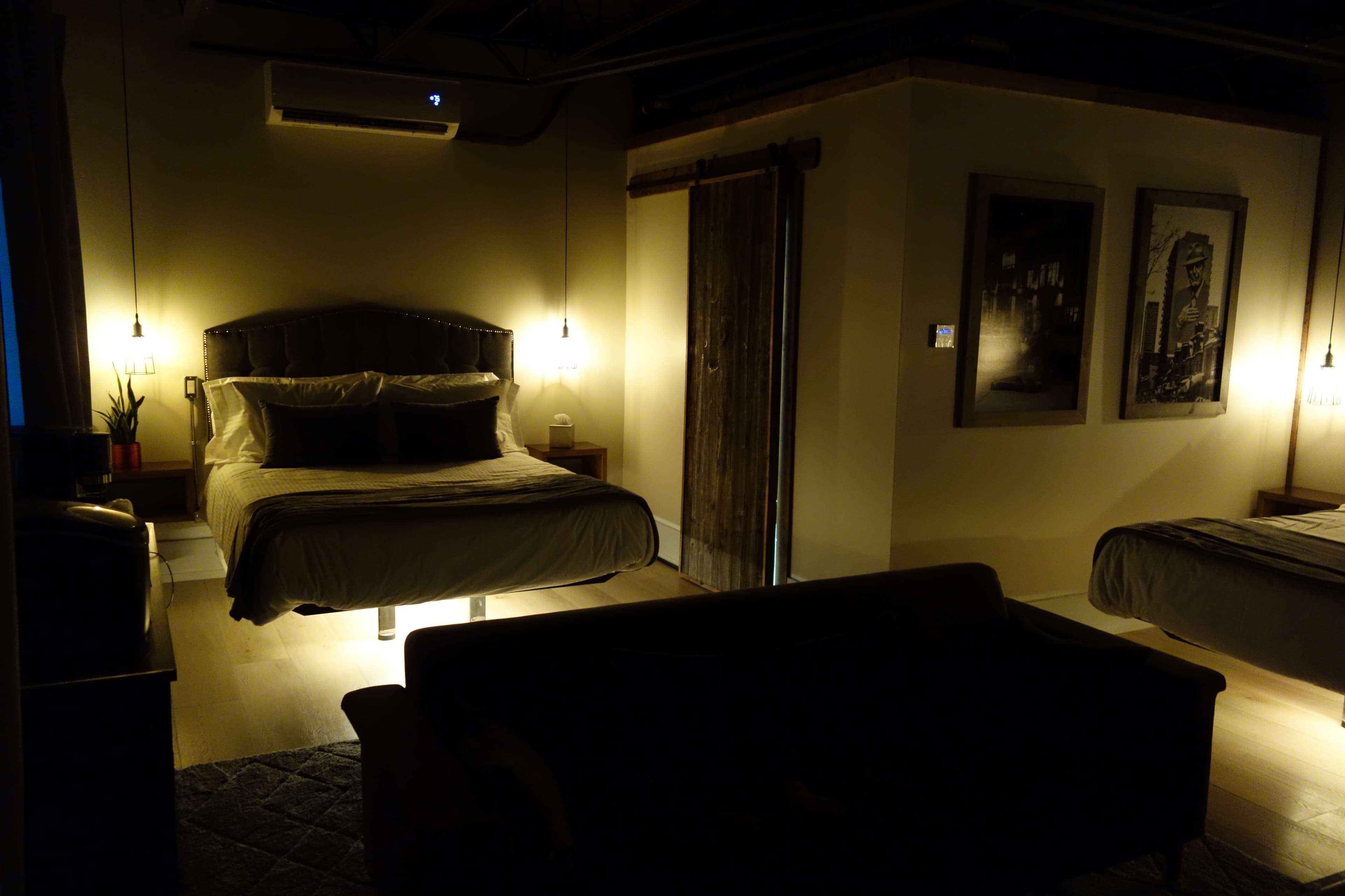 A dimly lit, moody industrial loft bedroom featuring two beds with under-lighting, a dark sofa, and a rustic sliding barn door.