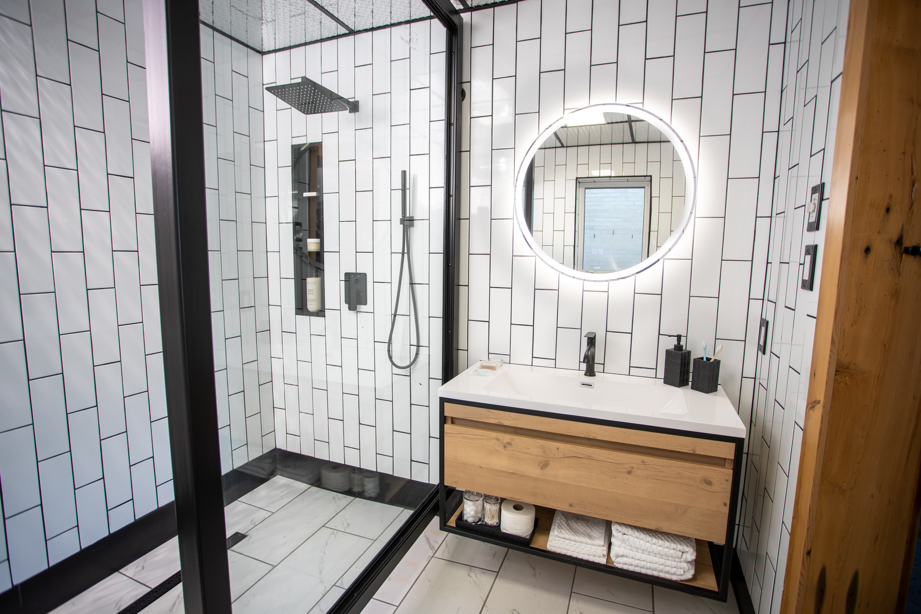 A stylish, modern bathroom with vertical white subway tiles, a black-framed walk-in shower, a wood vanity, and a backlit round mirror.
