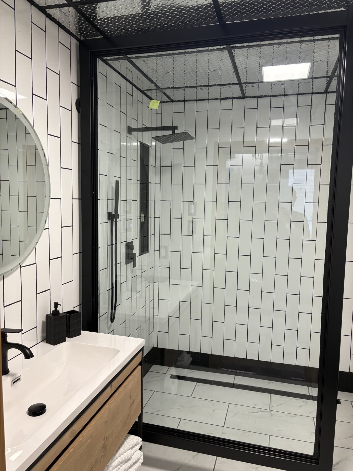A sleek, modern bathroom featuring a black-framed walk-in shower with white vertical subway tiles, a light wood vanity, and a round mirror.