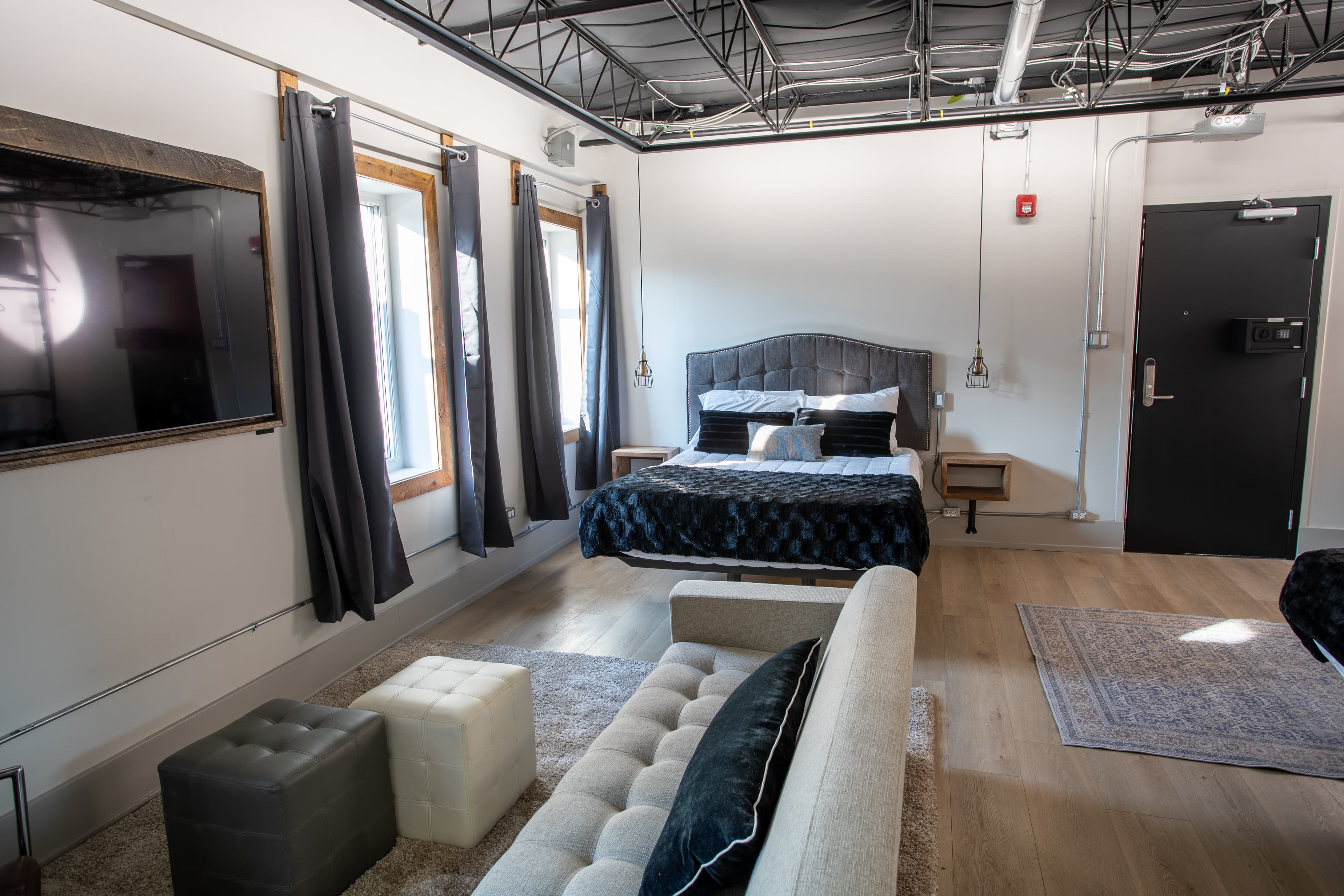 A chic industrial loft bedroom with a dark, tufted bed, a white tufted bench, a large mounted TV, and an exposed black ceiling.