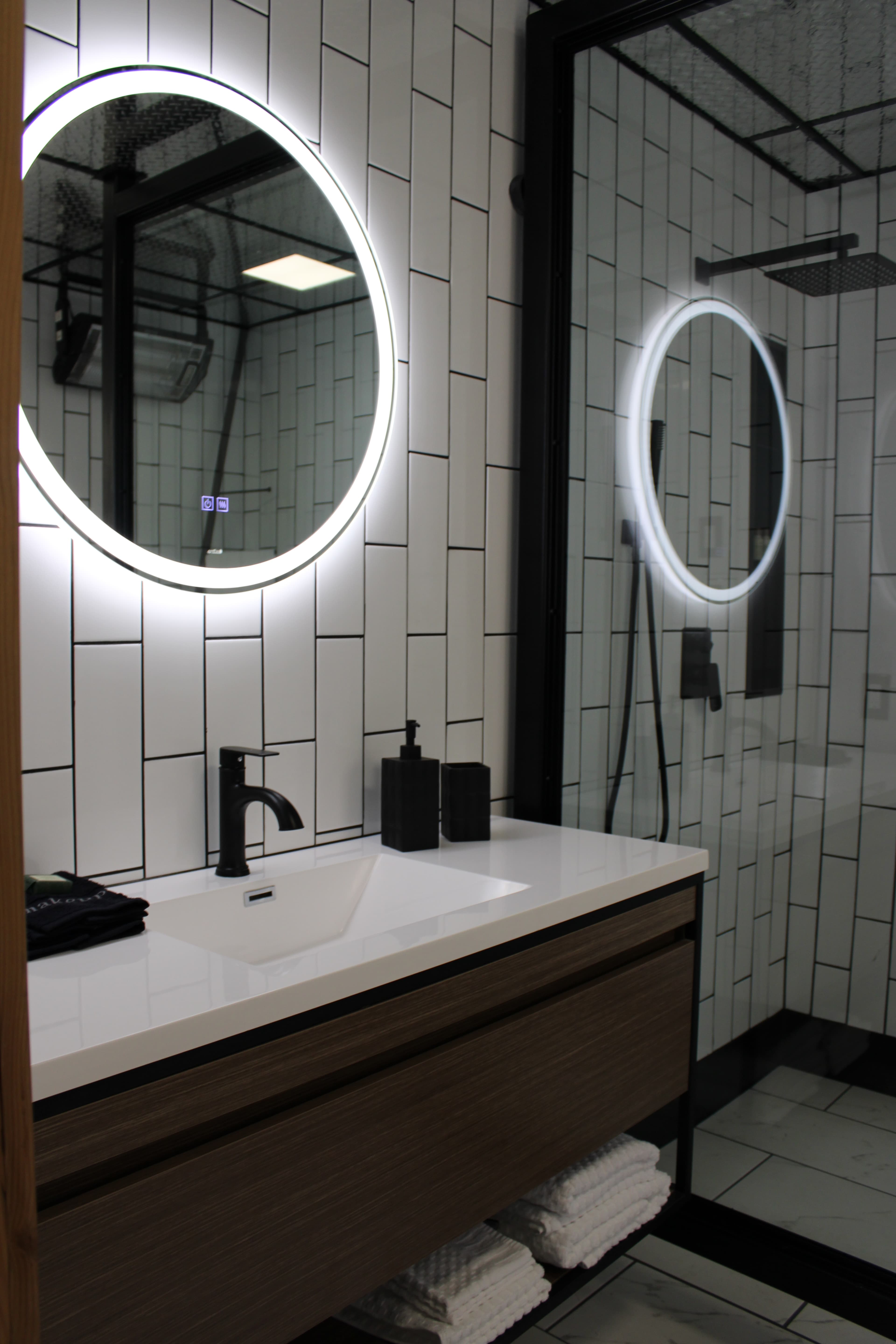 A stylish modern bathroom with a floating wood vanity, a backlit round mirror, and a sleek, black-framed walk-in shower.