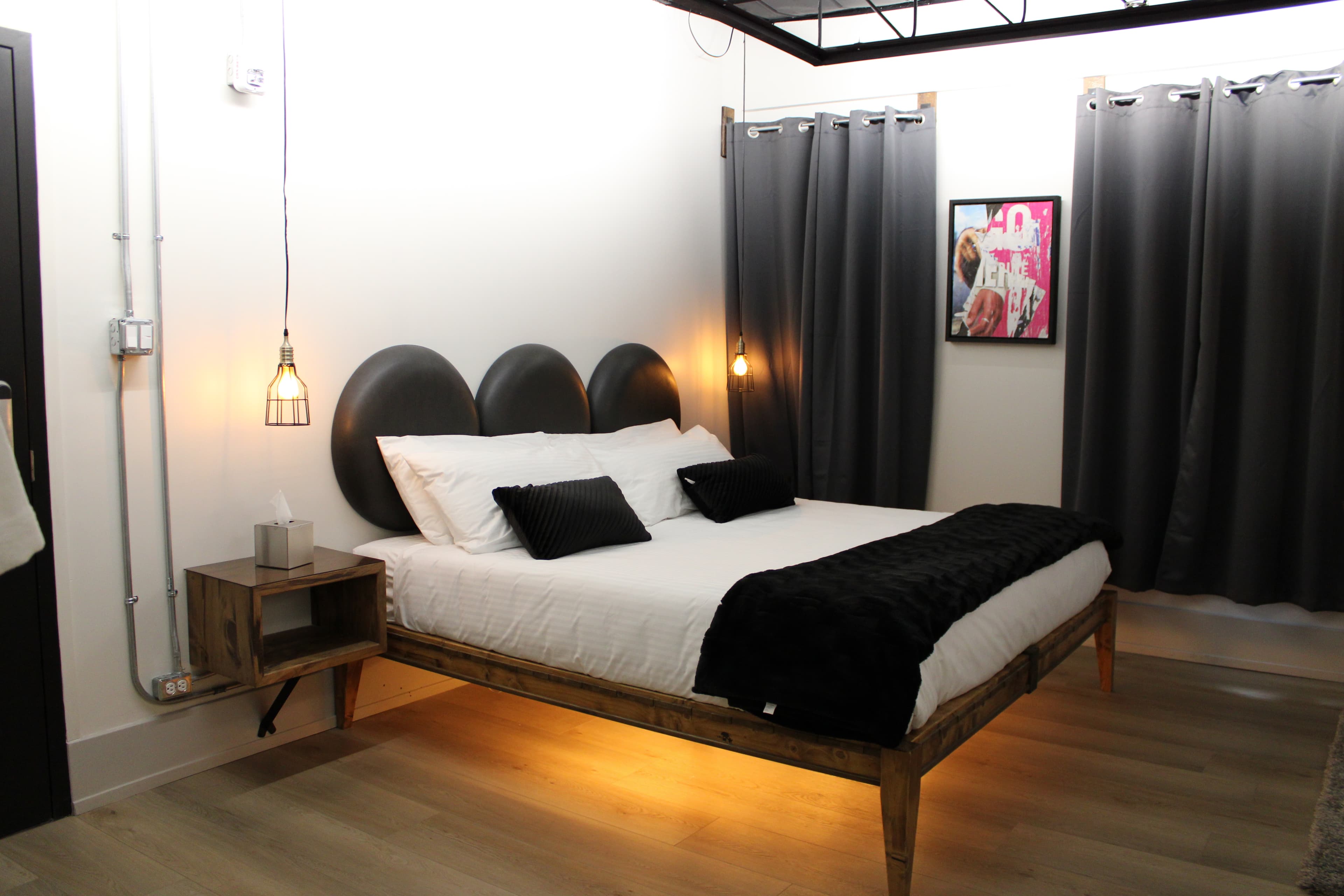 A trendy industrial loft bedroom with a unique leather headboard, wood bed frame with under-lighting, black bedding, and dark curtains.