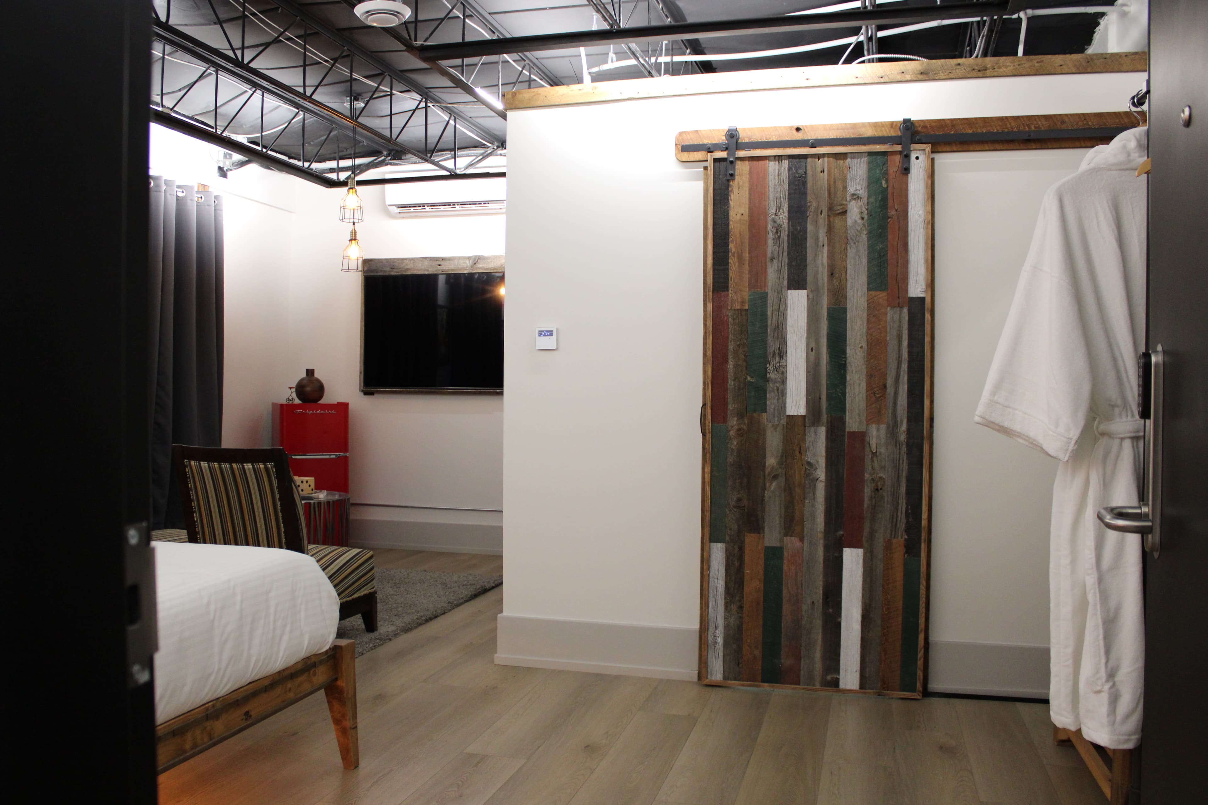 A stylish industrial-loft room with a rustic reclaimed wood sliding barn door, a plush white robe hanging up, and a view into the lounge area.