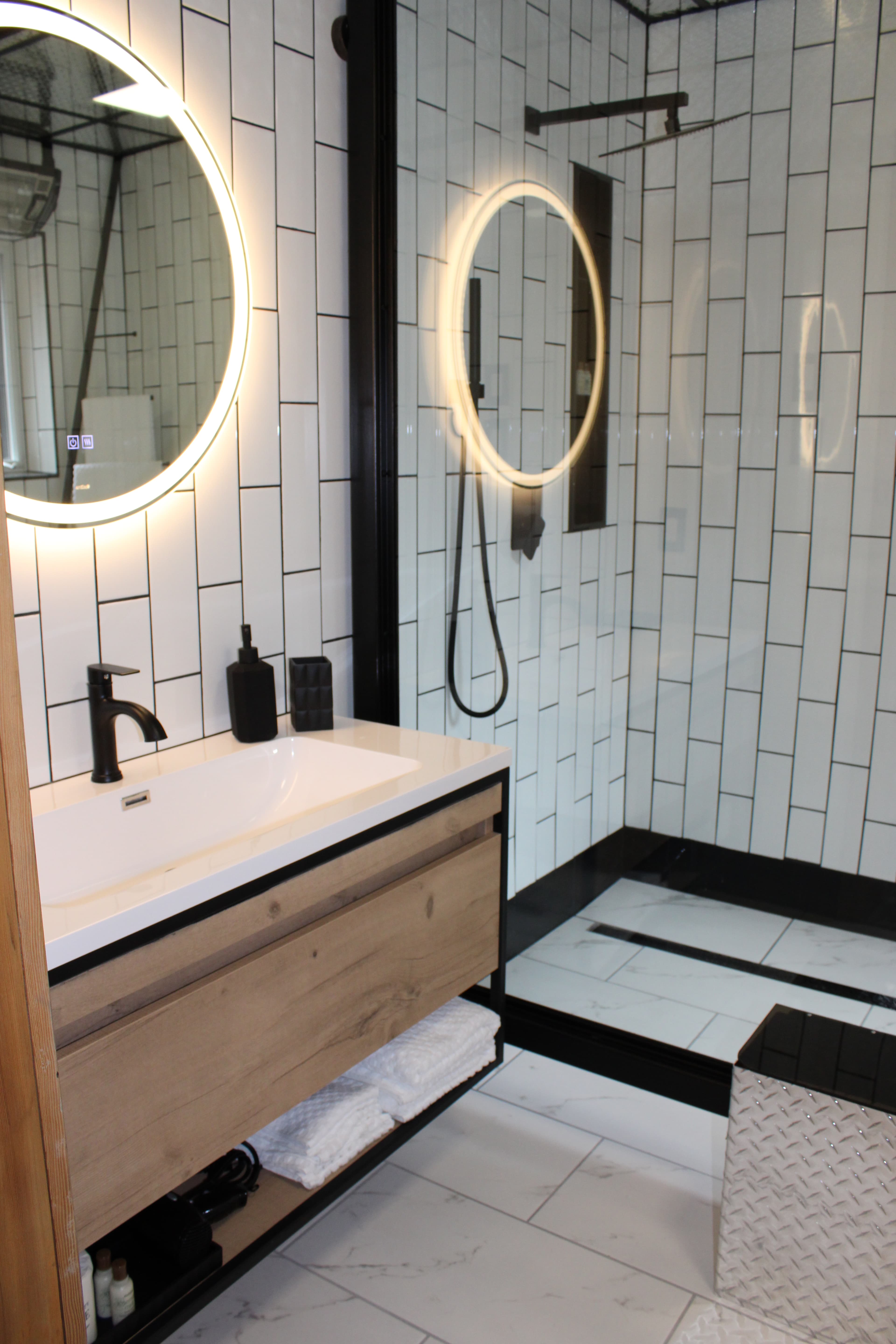 A stylish modern bathroom with a floating wood vanity, a backlit round mirror, and a sleek, black-framed walk-in shower with subway tiles.