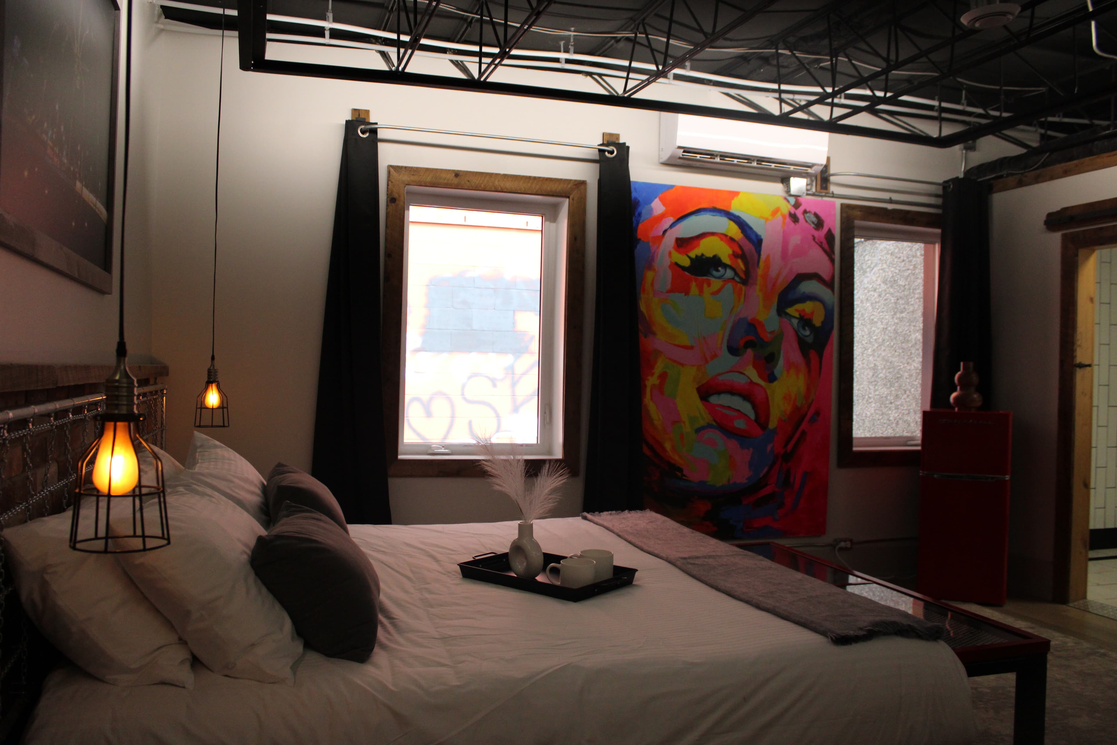 A bold industrial-style bedroom with a wood bed, a large, vibrant Marilyn Monroe pop art canvas, and a black exposed ceiling.
