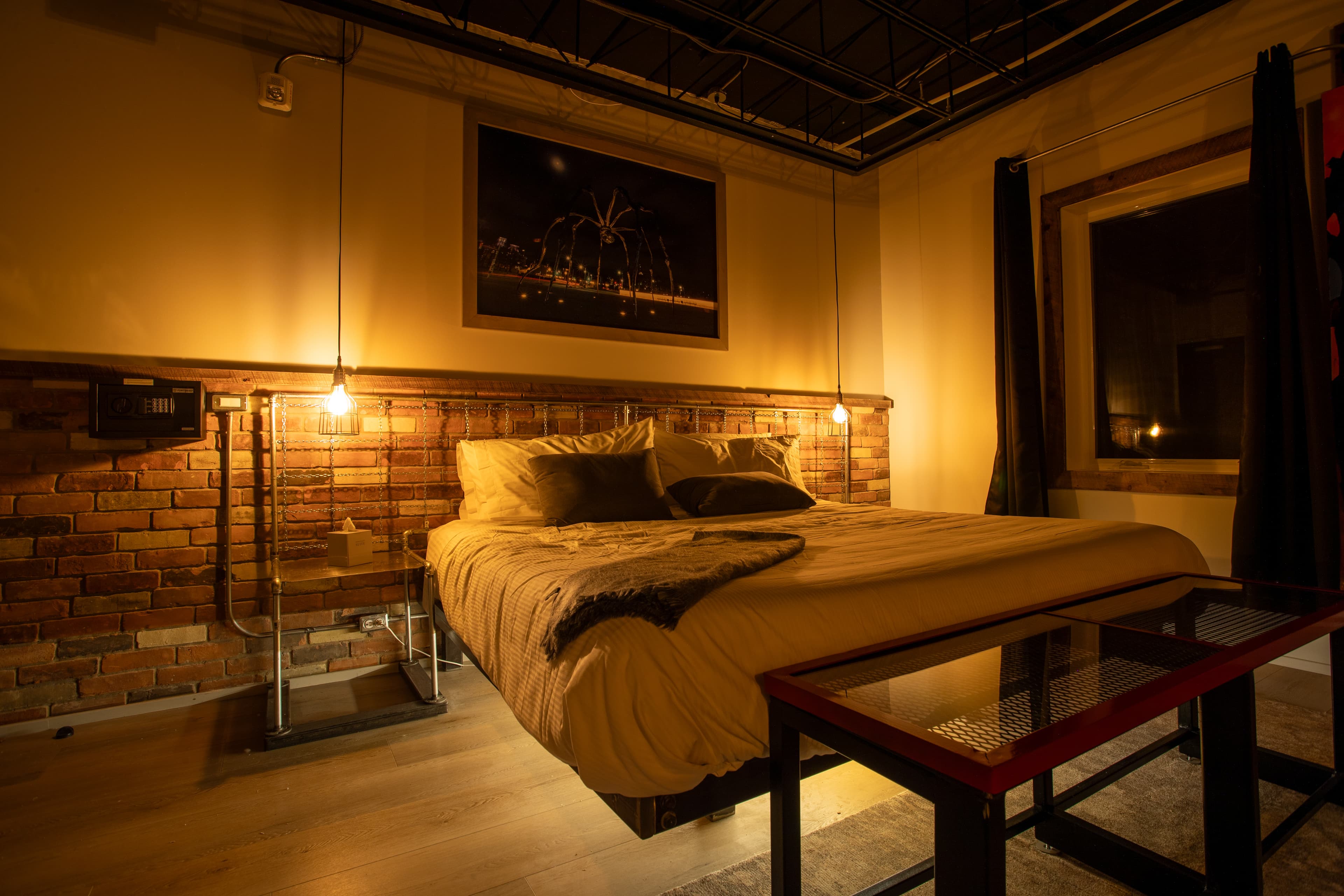 A moody, industrial loft bedroom featuring a brick accent wall, a low-profile bed with under-lighting, and warm hanging Edison bulb lights.