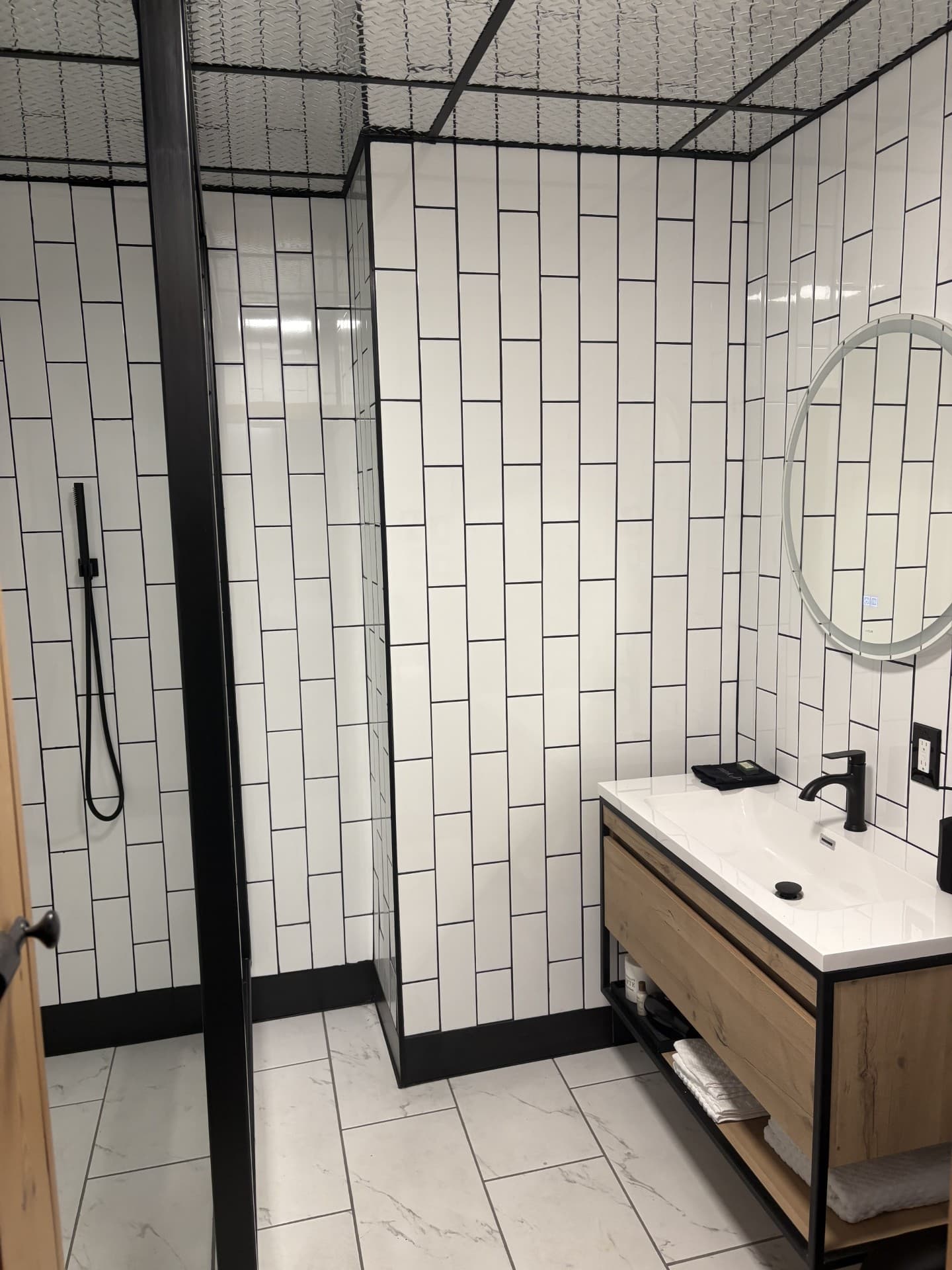 A chic, modern bathroom featuring a black-framed walk-in shower and a light wood floating vanity with a round mirror and subway tiles.