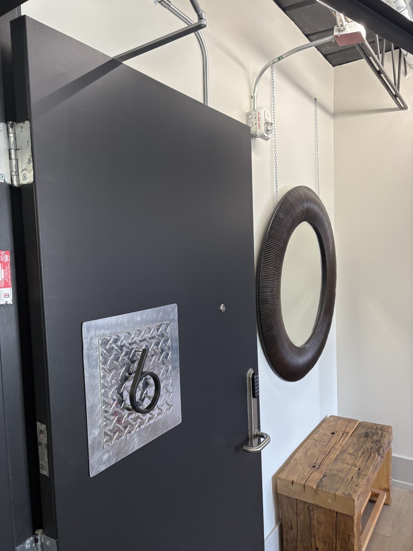 The entryway of a loft unit, featuring a dark door with a large, decorative "6" house number, a round mirror, and a wooden bench.