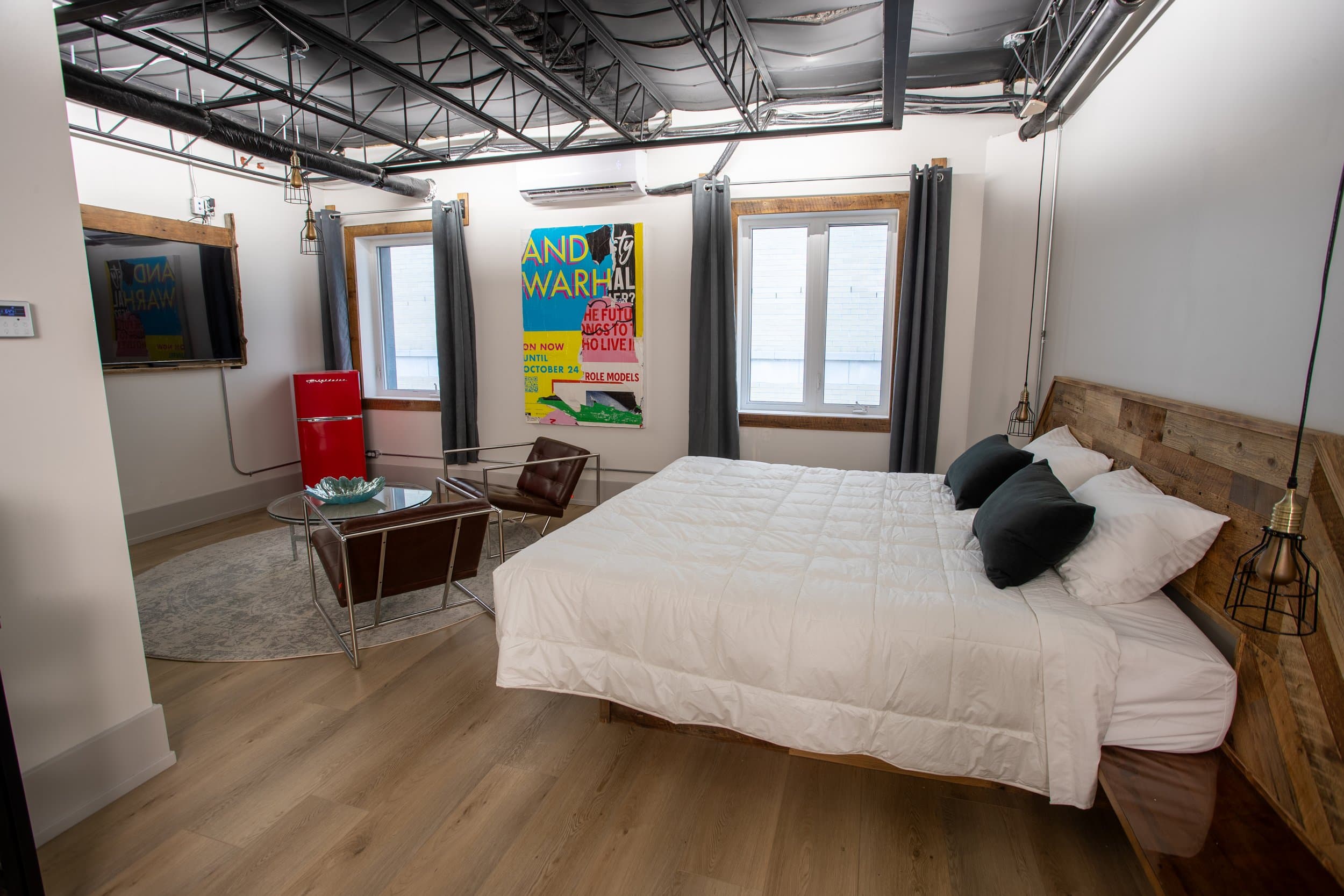 A spacious, stylish hotel room featuring a king bed, exposed black ceiling, modern artwork, wood accents, and a retro red mini-fridge.