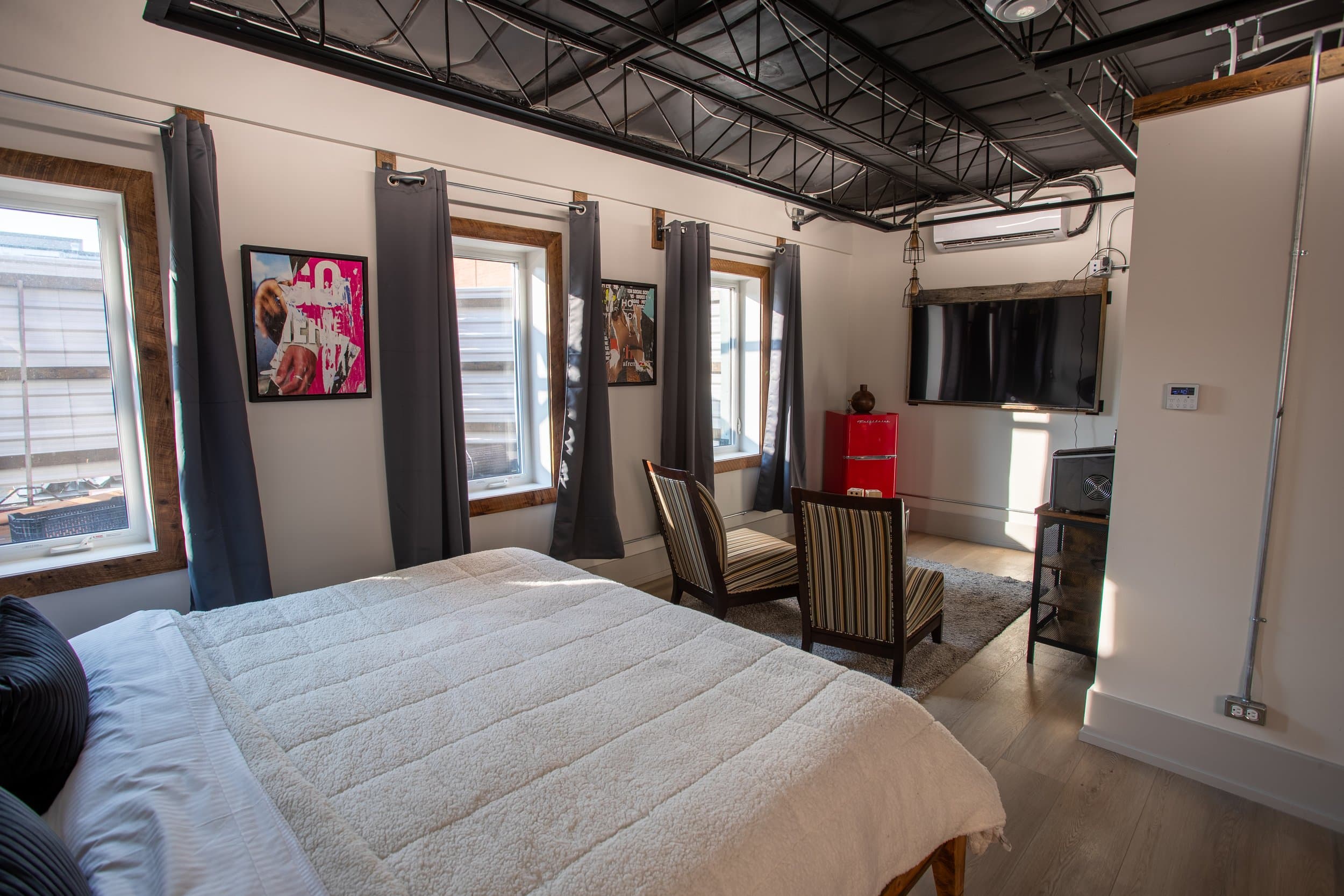 A bright, modern hotel room with a king bed, exposed black ceiling, multiple windows, a seating area, and pop art on the walls.
