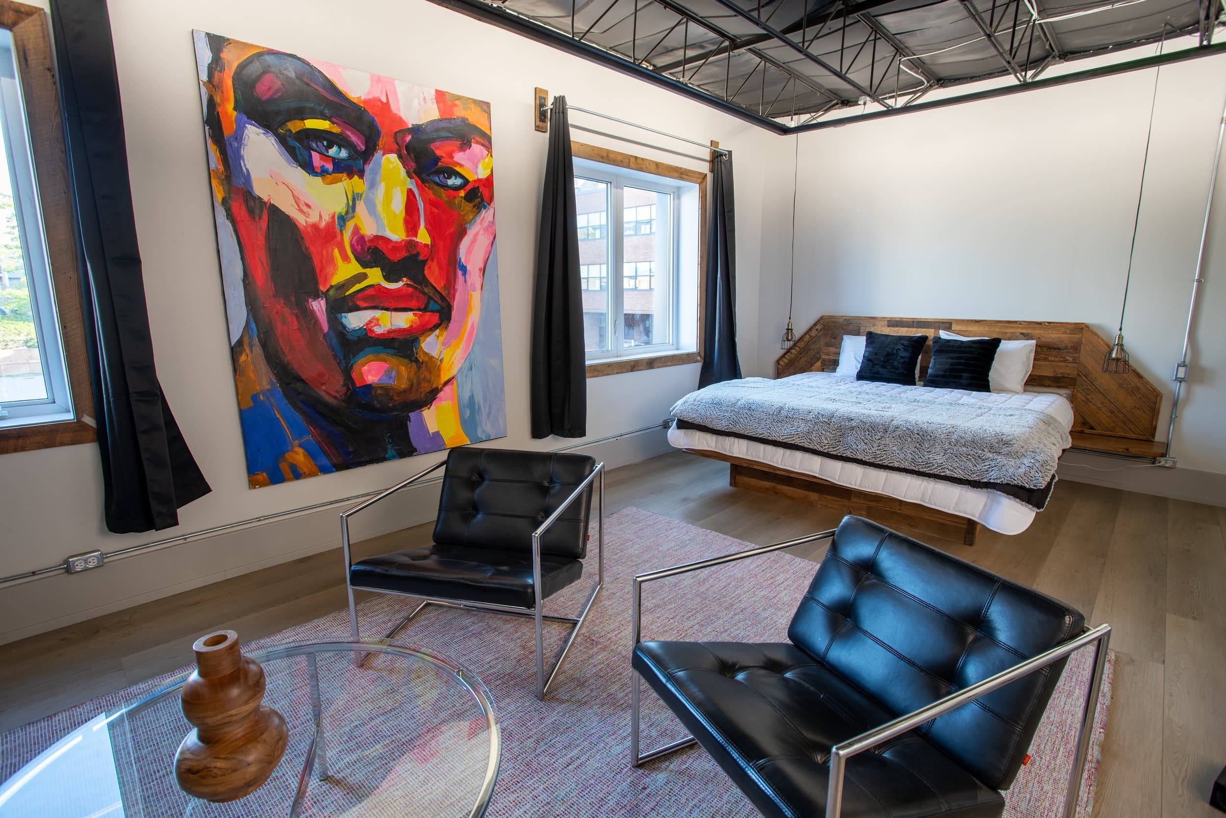 A striking, modern hotel room with a king platform bed, a pair of black tufted chairs, and a large, colorful pop art portrait on the wall.