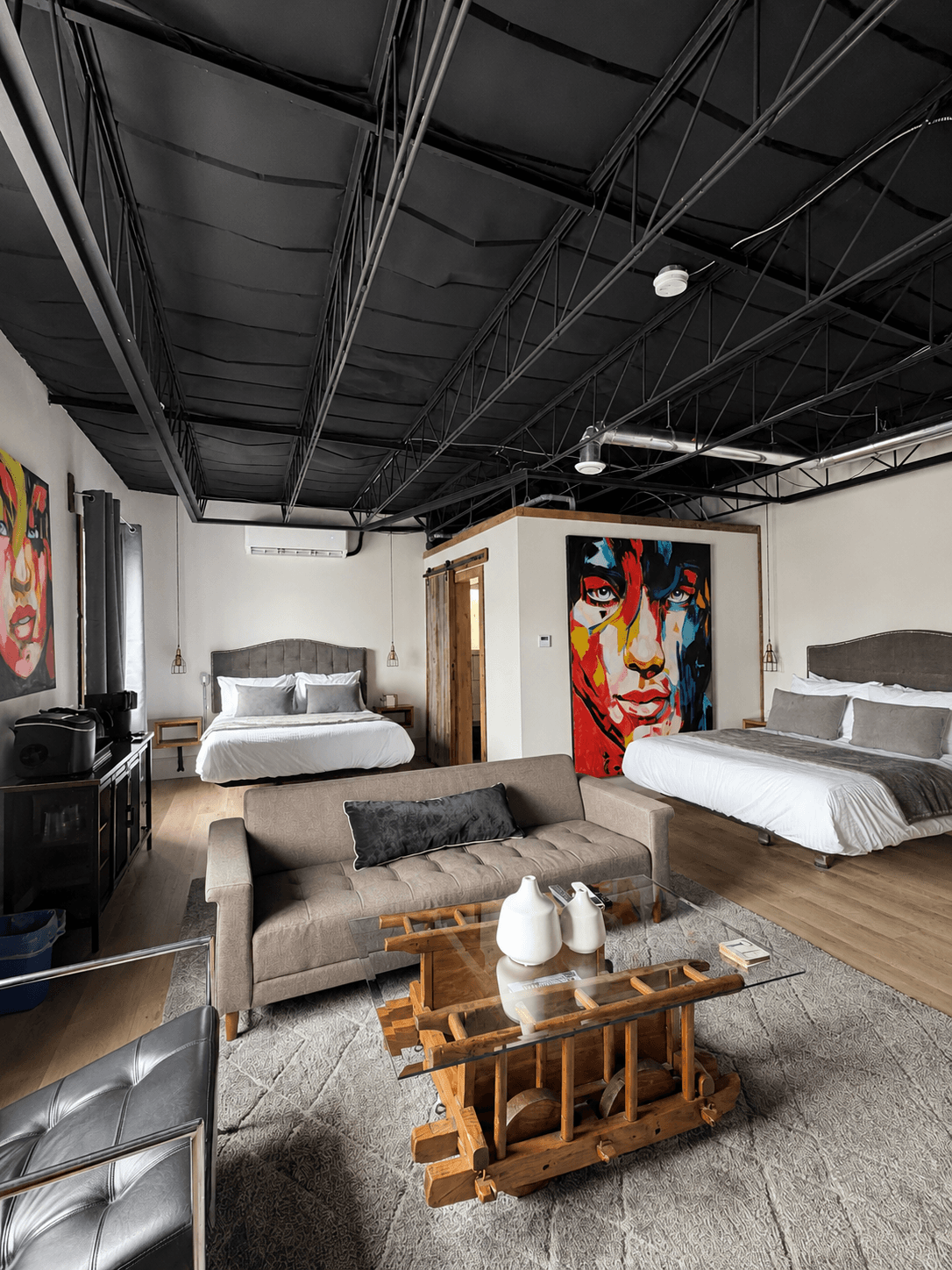 A modern bedroom featuring two beds, a plush sofa, artistic wall murals, and natural wood accents.