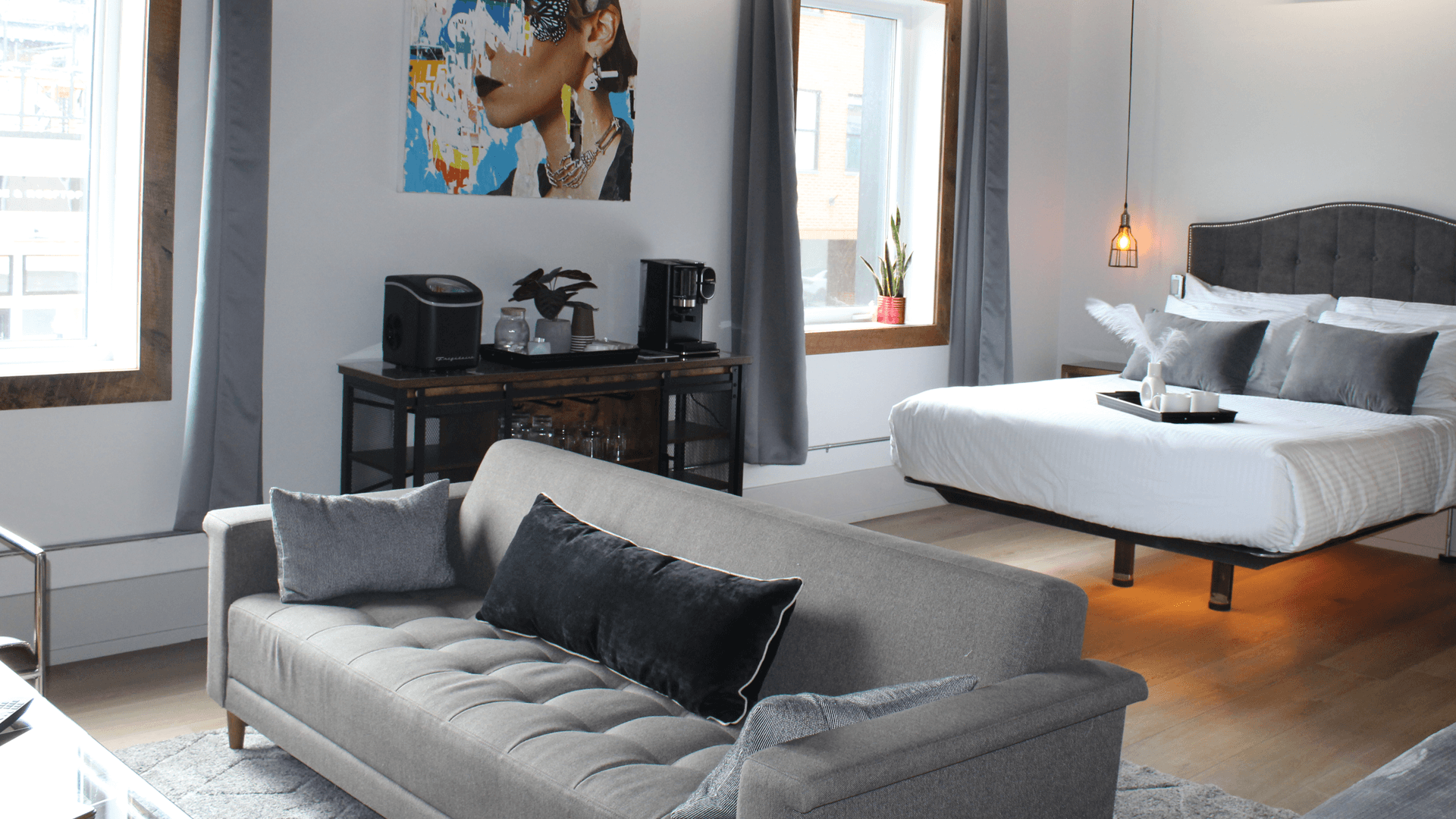 Modern hotel room with a grey sofa, black velvet cushions, and a bed. A vibrant portrait hangs above a coffee bar. Bright windows and plants add a cozy feel.
