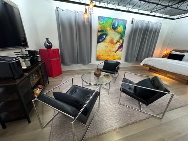 A modern living space featuring a glass table with black chairs, colorful artwork, and a cozy bed in the background.