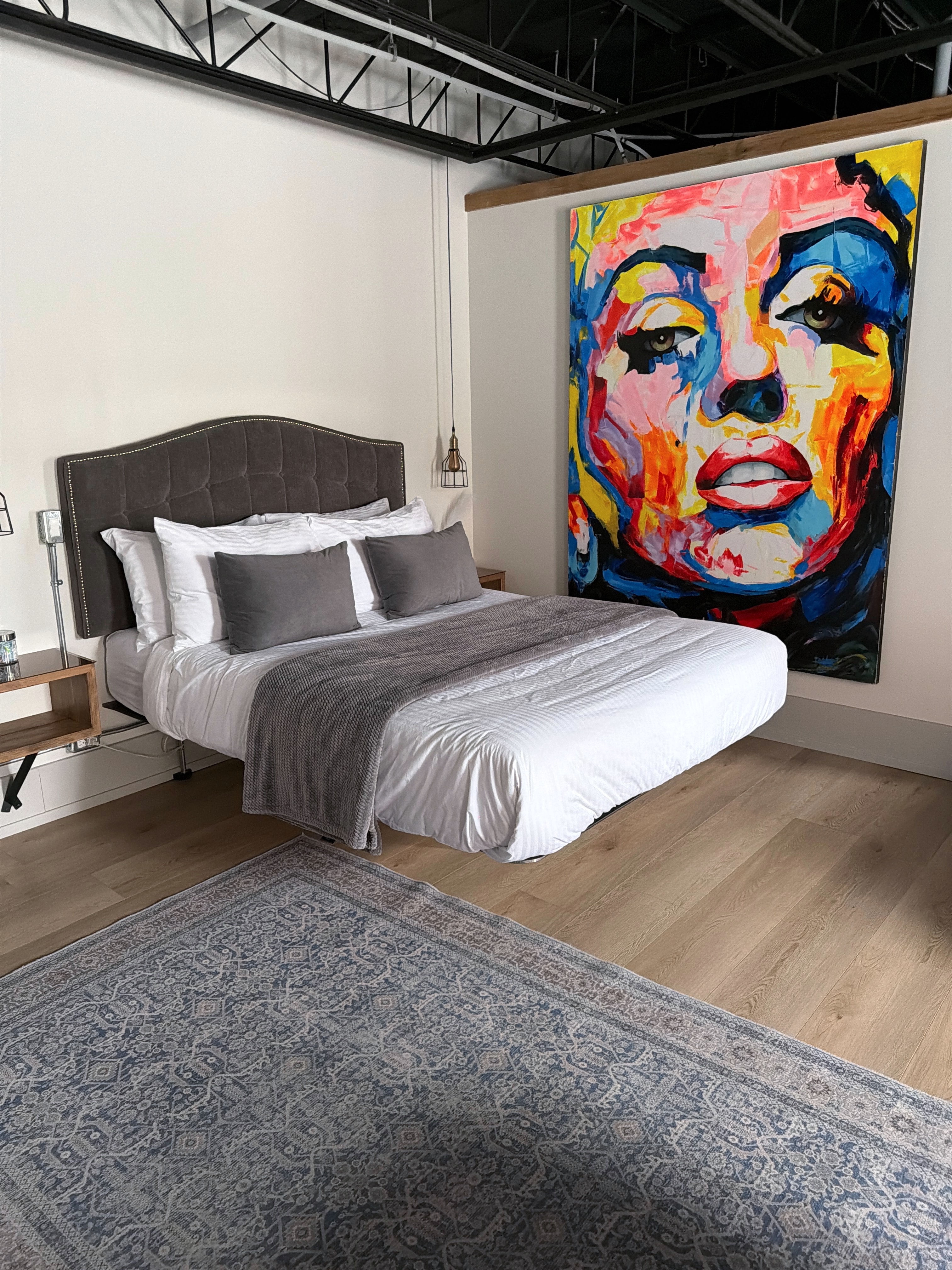 A stylish bedroom featuring a bed with gray and white bedding, accent pillows, and a vibrant portrait on the wall.