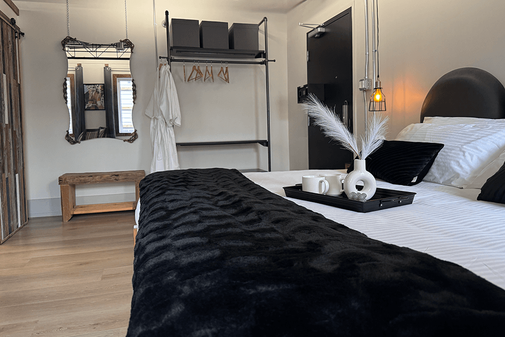 A stylish, modern bedroom featuring a bed with black throw blankets, a wooden bench, a mirror, and decorative items on a tray.