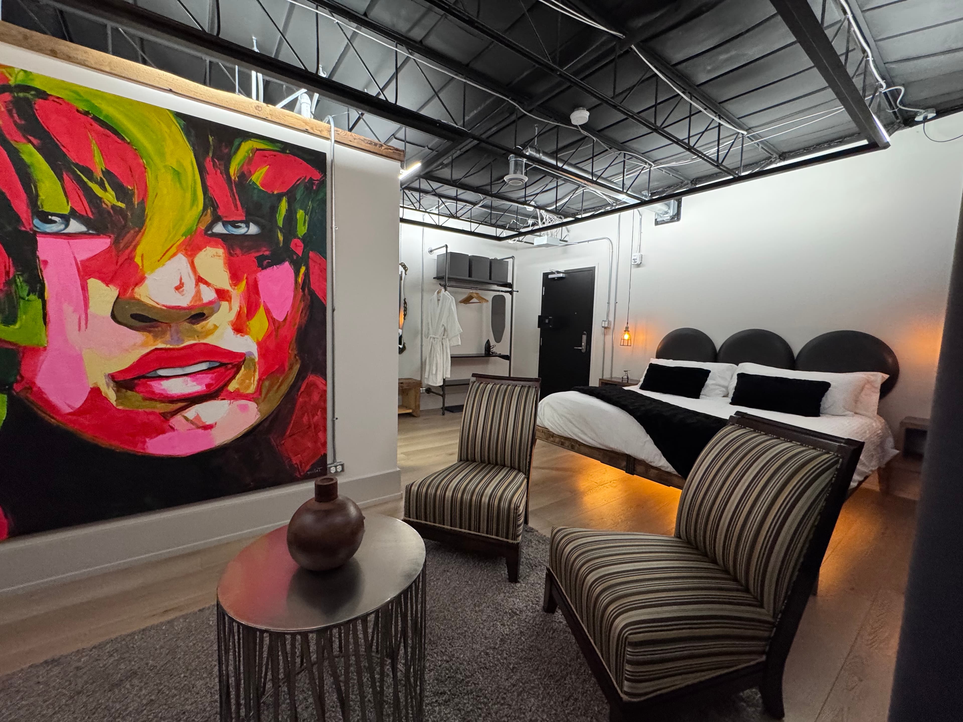 A modern bedroom featuring a colorful abstract portrait, two striped chairs, and a stylish metal side table.