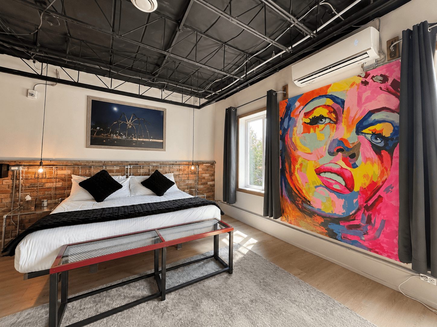 A stylish bedroom featuring a bed with black and white linens, a large colorful mural of a woman, and modern decor.