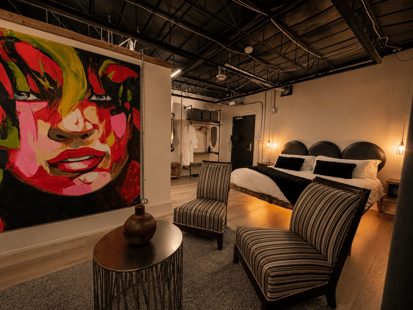 A stylish hotel room featuring a colorful mural, two striped chairs, and a bed with dark bedding.