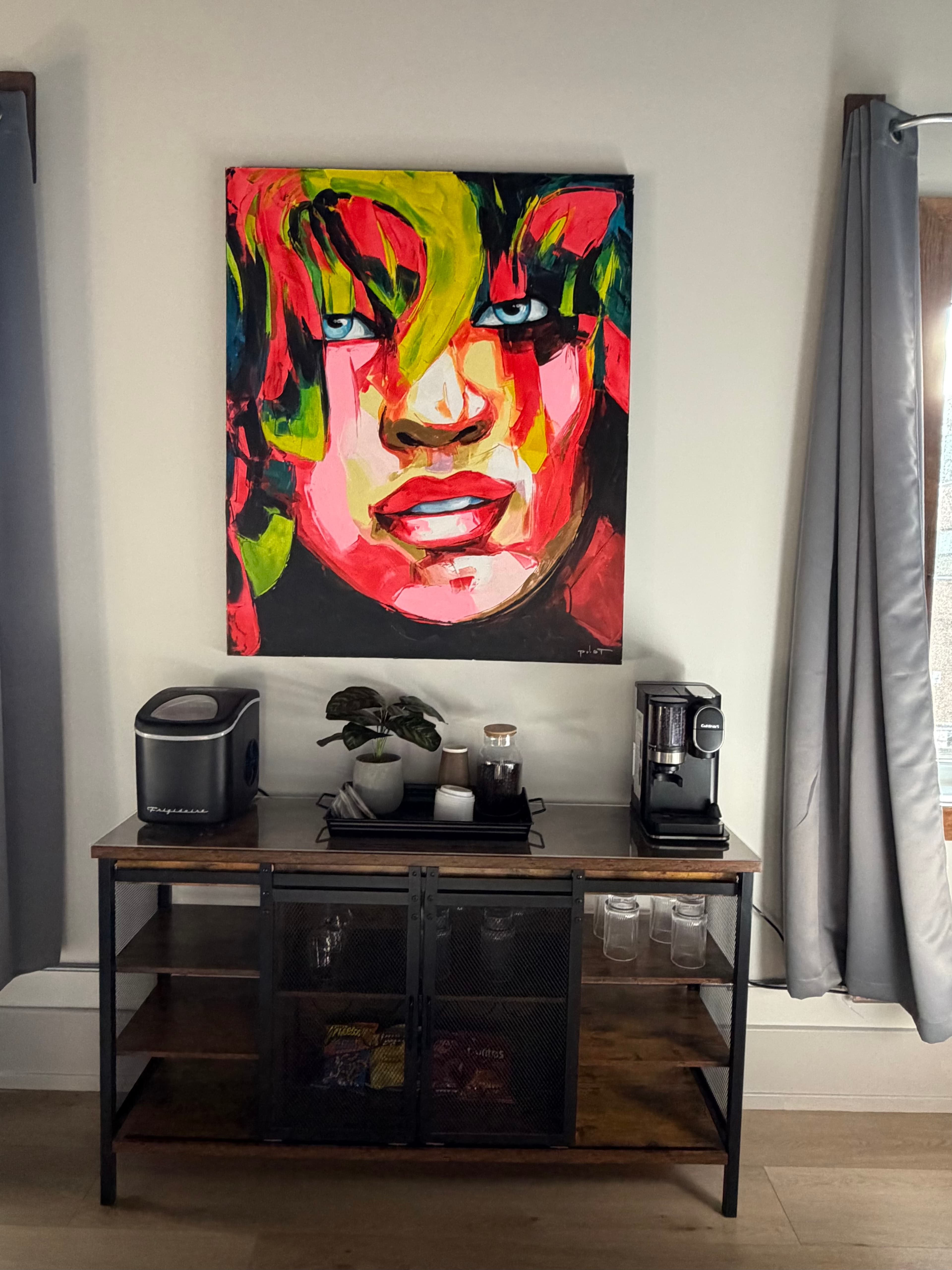 A colorful portrait of a woman hangs above a sideboard with a coffee maker and various accessories.