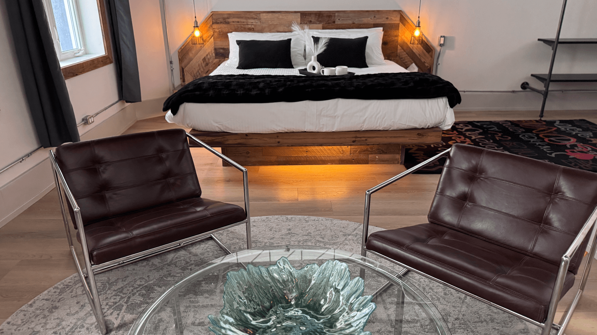 A modern bedroom with a wooden bed, white linens, and black accents. Two leather chairs face a glass table in the foreground, creating a cozy, stylish atmosphere.