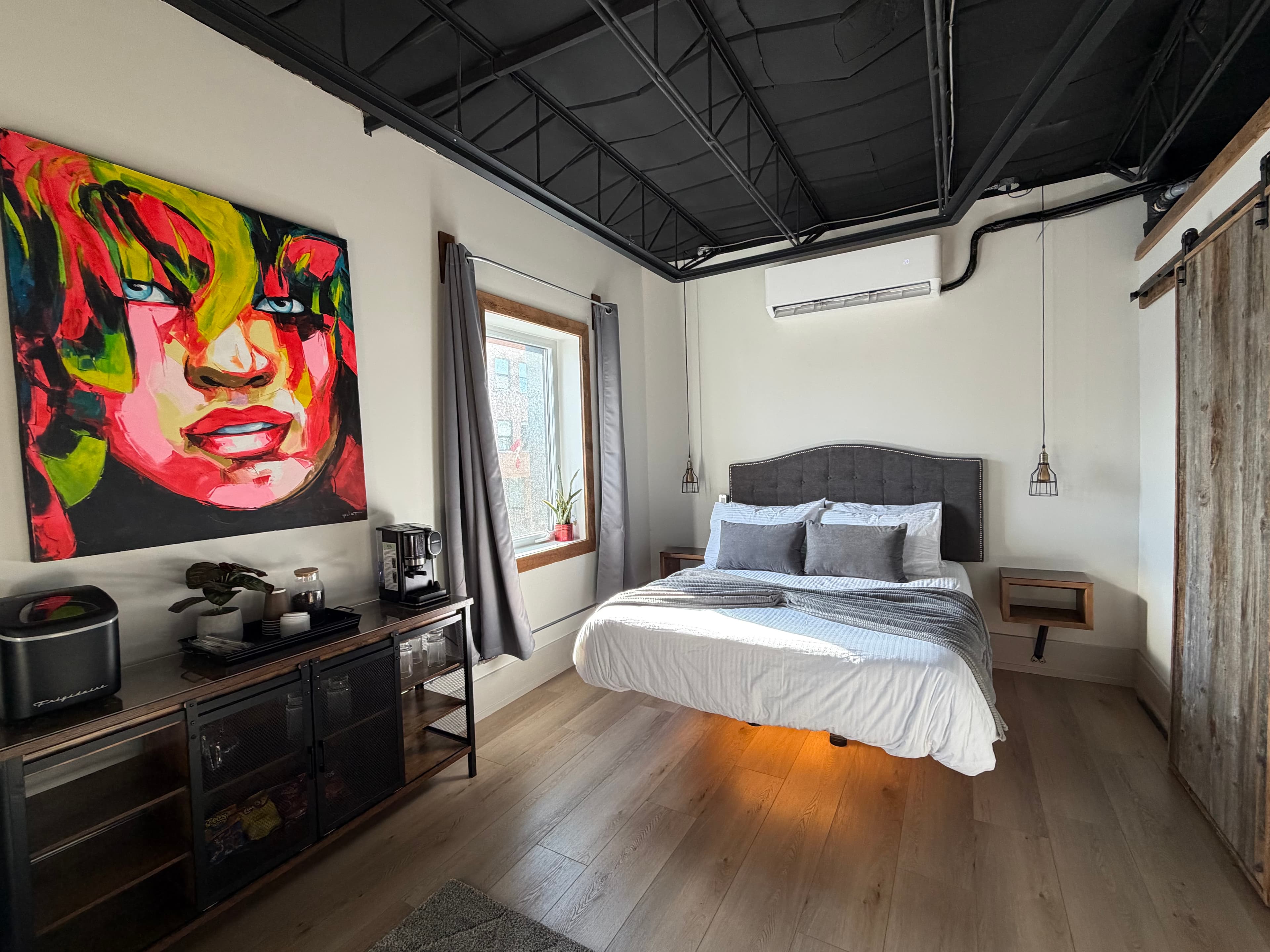 A spacious, modern industrial loft with two beds, a gray sofa, and a rustic sliding barn door leading to a bathroom.