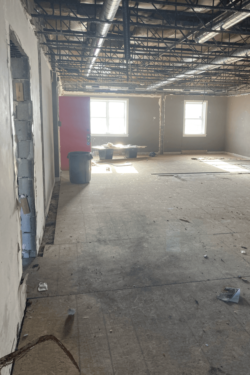 Empty room under construction with exposed ceiling ducts, a red door, and two windows letting in sunlight. Debris is scattered on the bare floor.