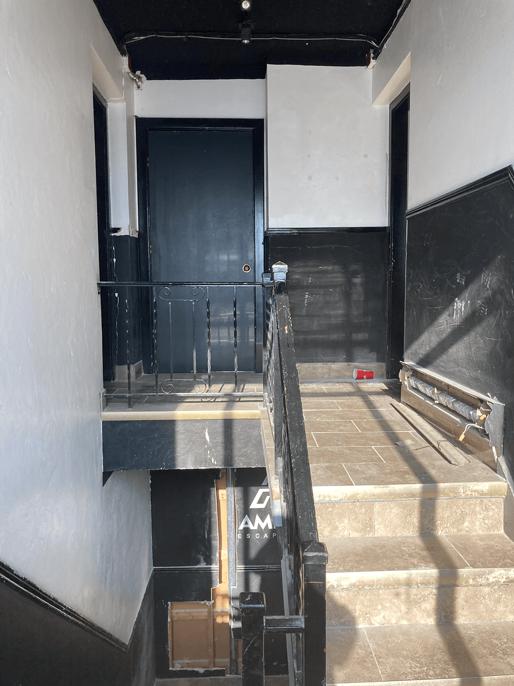 Dimly lit hallway with black doors and handrails, white walls, and tiled floors. Sunlight casts shadows on the stairs, creating a stark, minimalist tone.