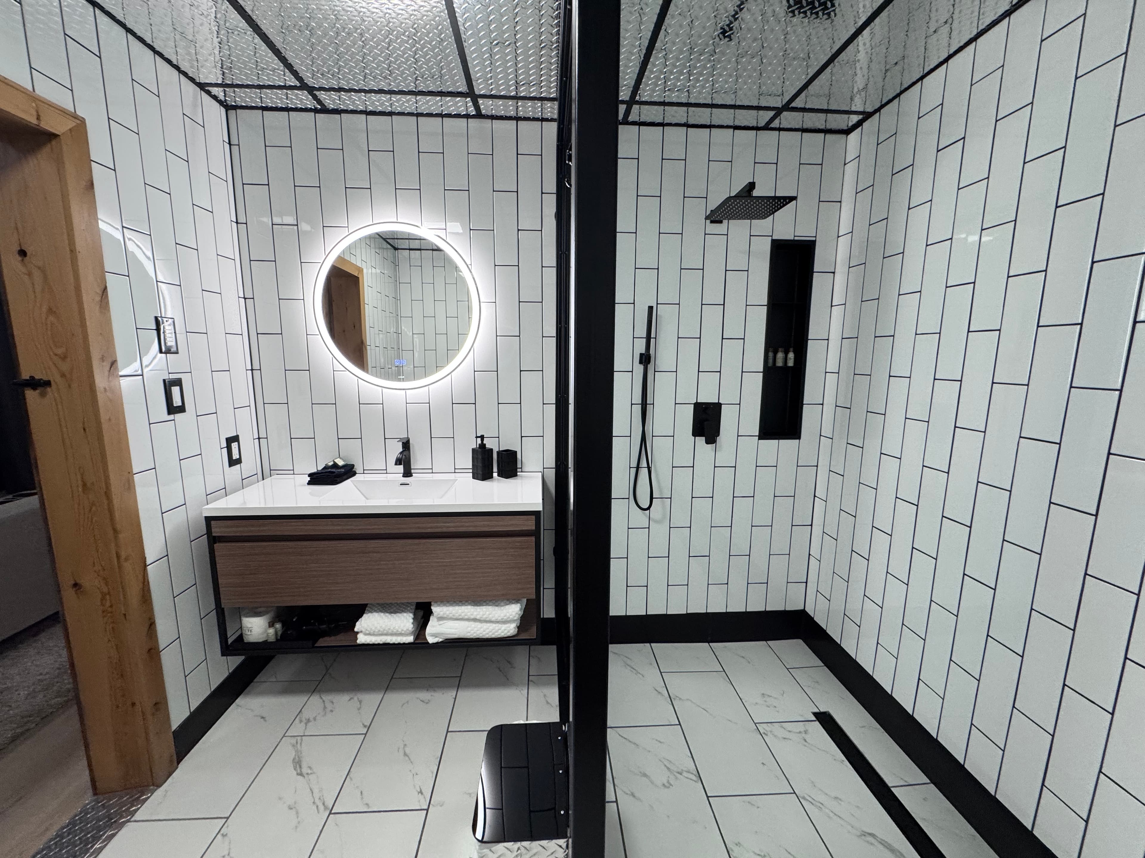 A modern bathroom featuring white tiled walls, a round illuminated mirror, and a sleek shower area.