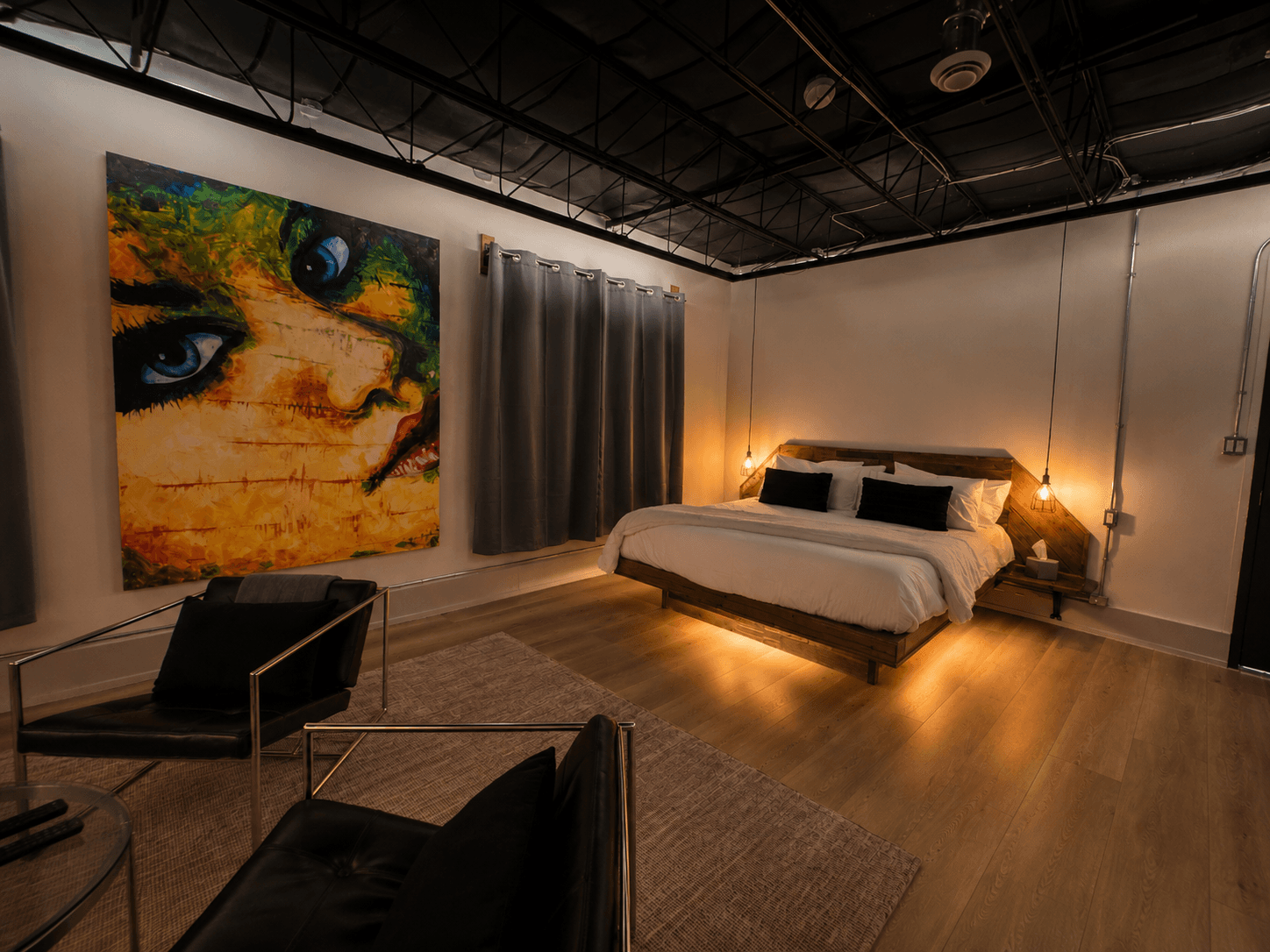 A modern bedroom featuring a striking wall art, a bed with white linens, gray curtains, and stylish furniture.