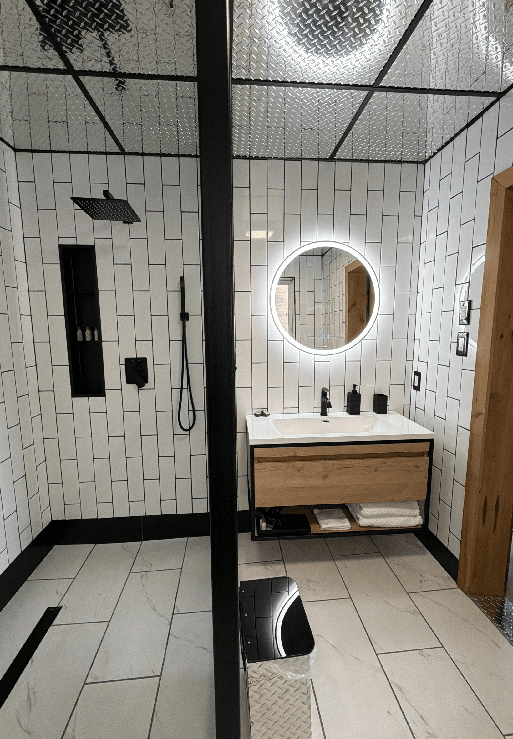 Modern bathroom featuring white tiled walls, a circular mirror with backlighting, a wooden vanity, and a sleek shower area.