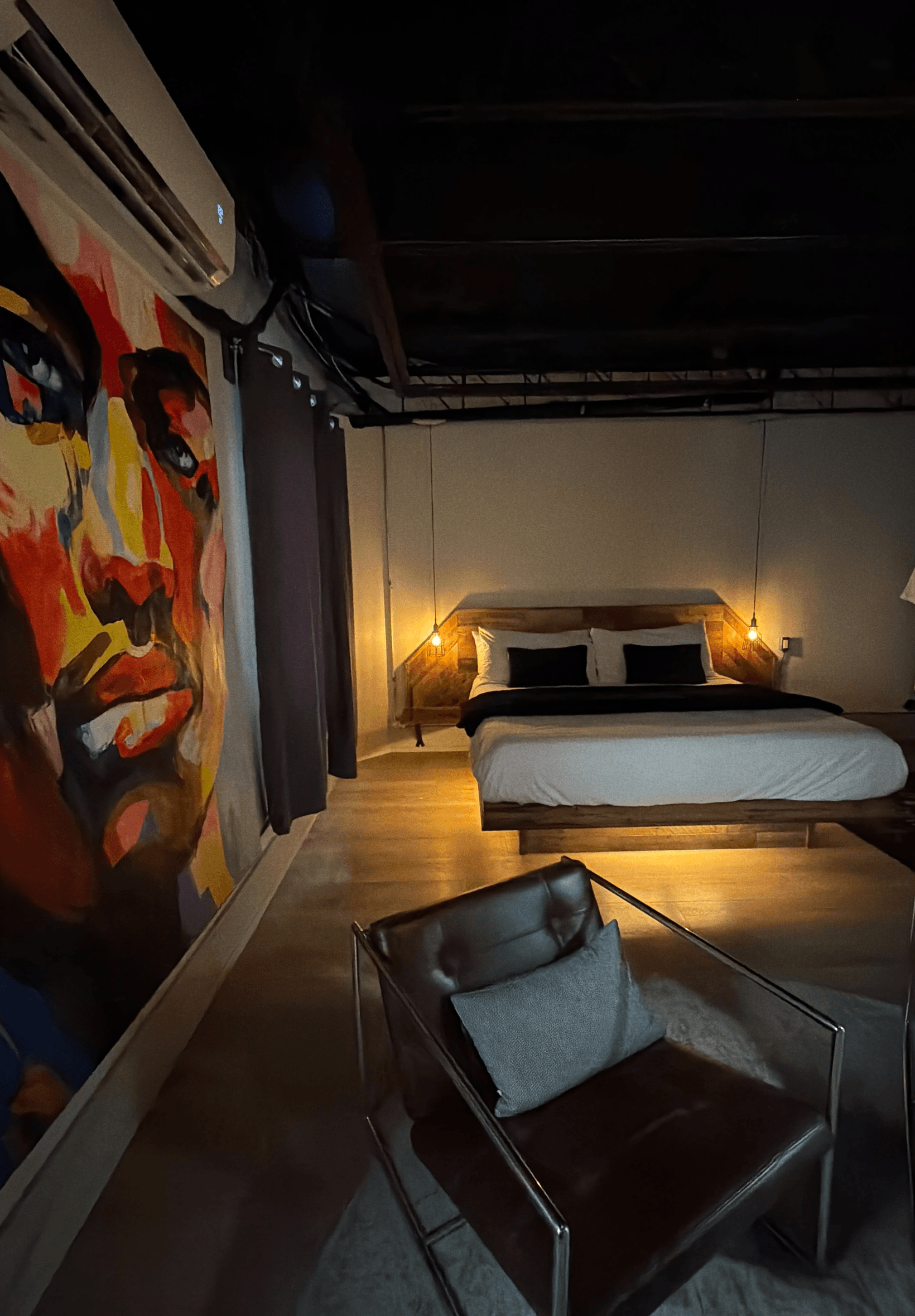 A modern bedroom featuring a large colorful mural, a wooden bed with black and white bedding, and a cozy armchair.